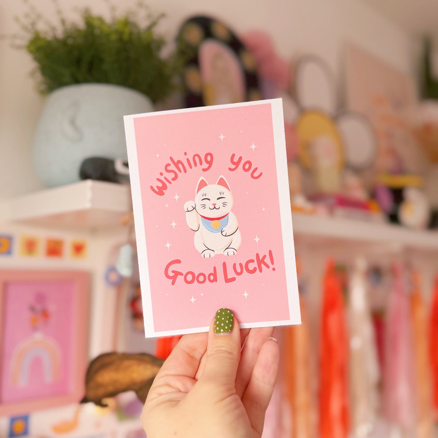 Hand holding a pink card with a cat illustration and 'Wishing you Good Luck!' text, blurred background with toys and decor.