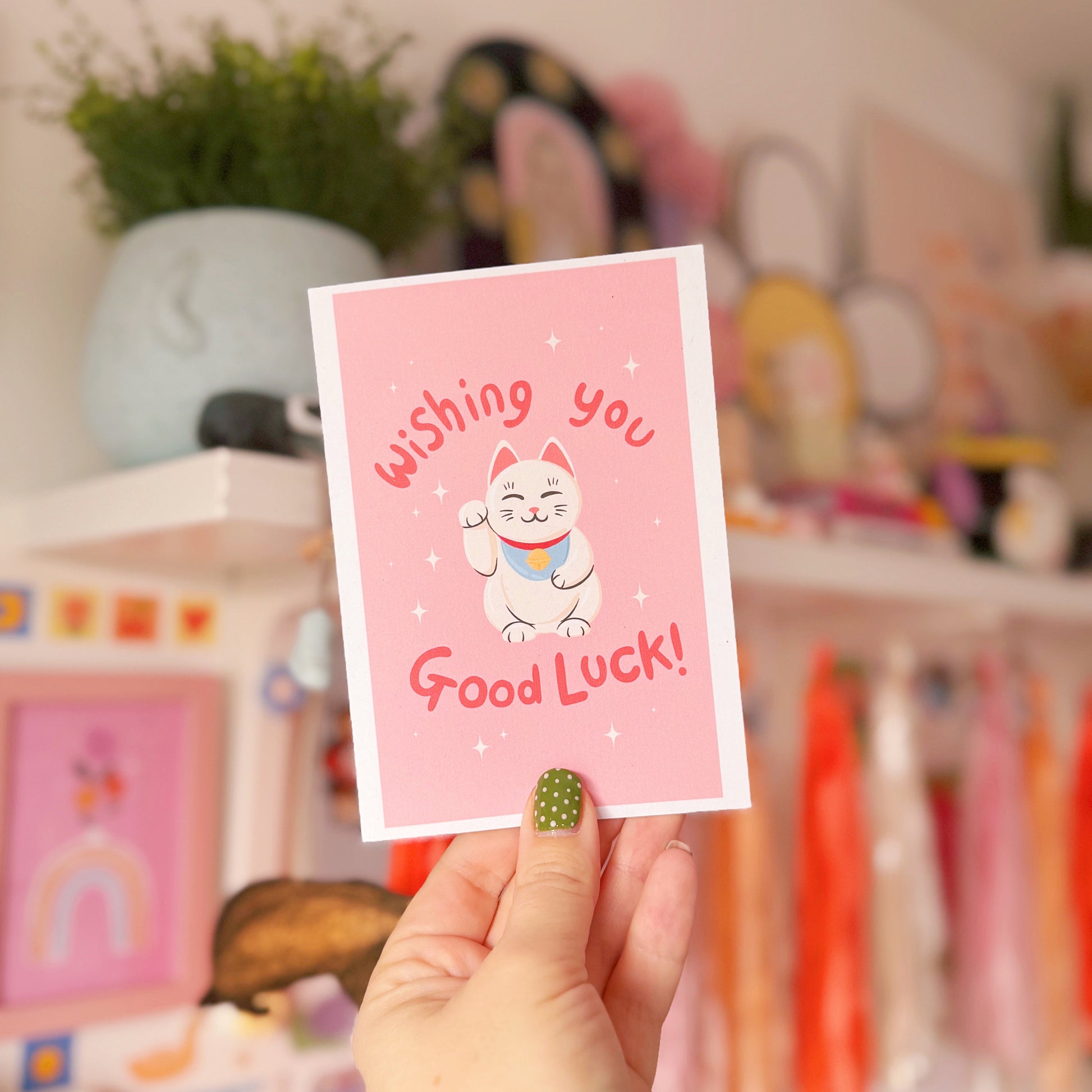 Hand holding a pink card with a cat illustration and 'Wishing you Good Luck!' text, blurred background with toys and decor.