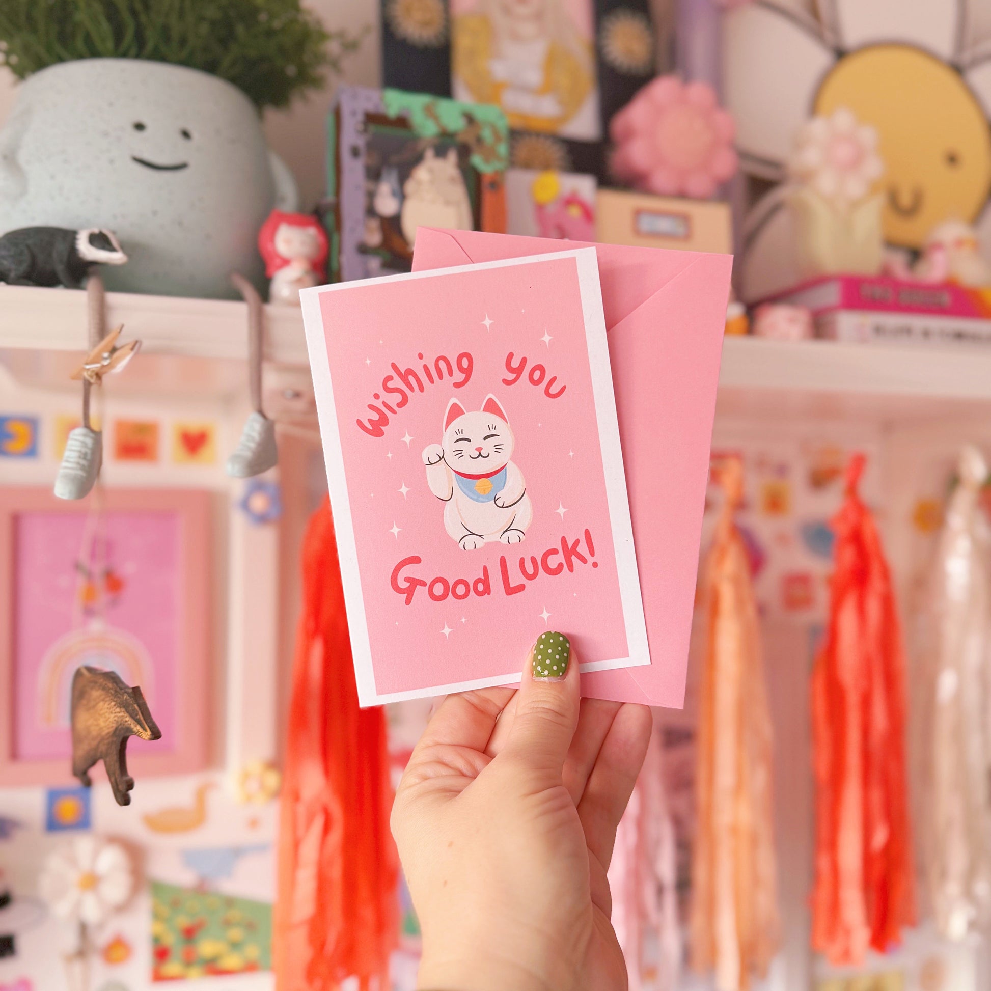 Hand holding a pink 'Good Luck' card with a cat illustration in a colourful room.