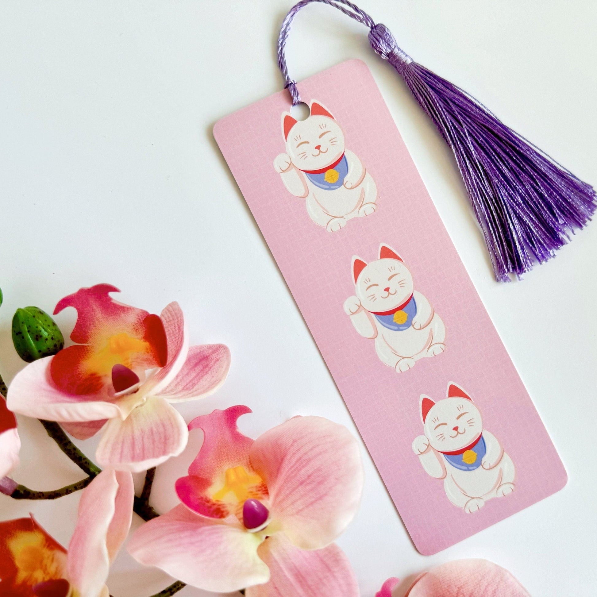 A picture of a pink bookmark, with a coordinating purple tassel, with three illustrations of Japanese lucky cats on the front.