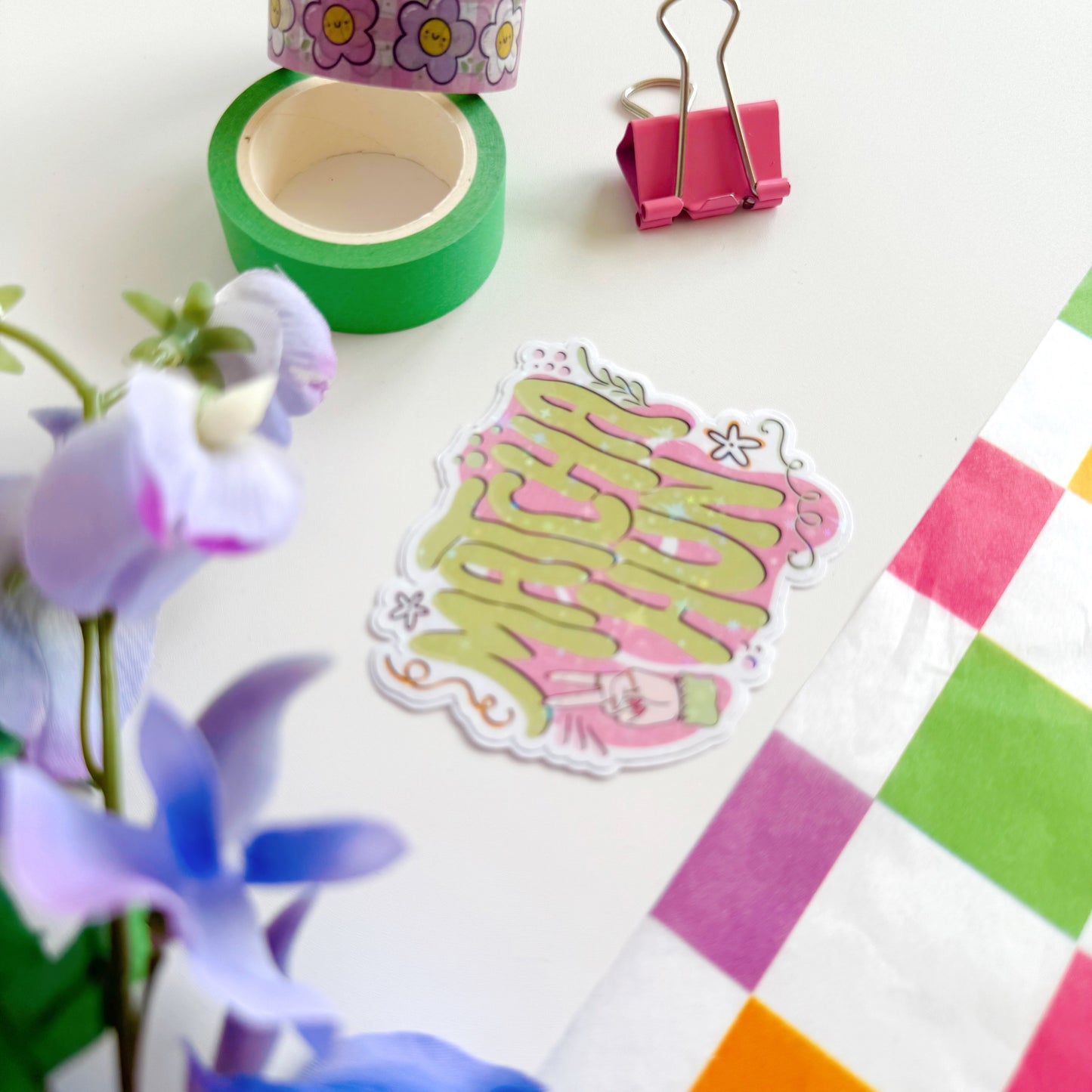 A holographic sticker with text saying 'Matcha Hun' in funky bubble writing and other hand illustrated elements around the text. The colours are pink and green.