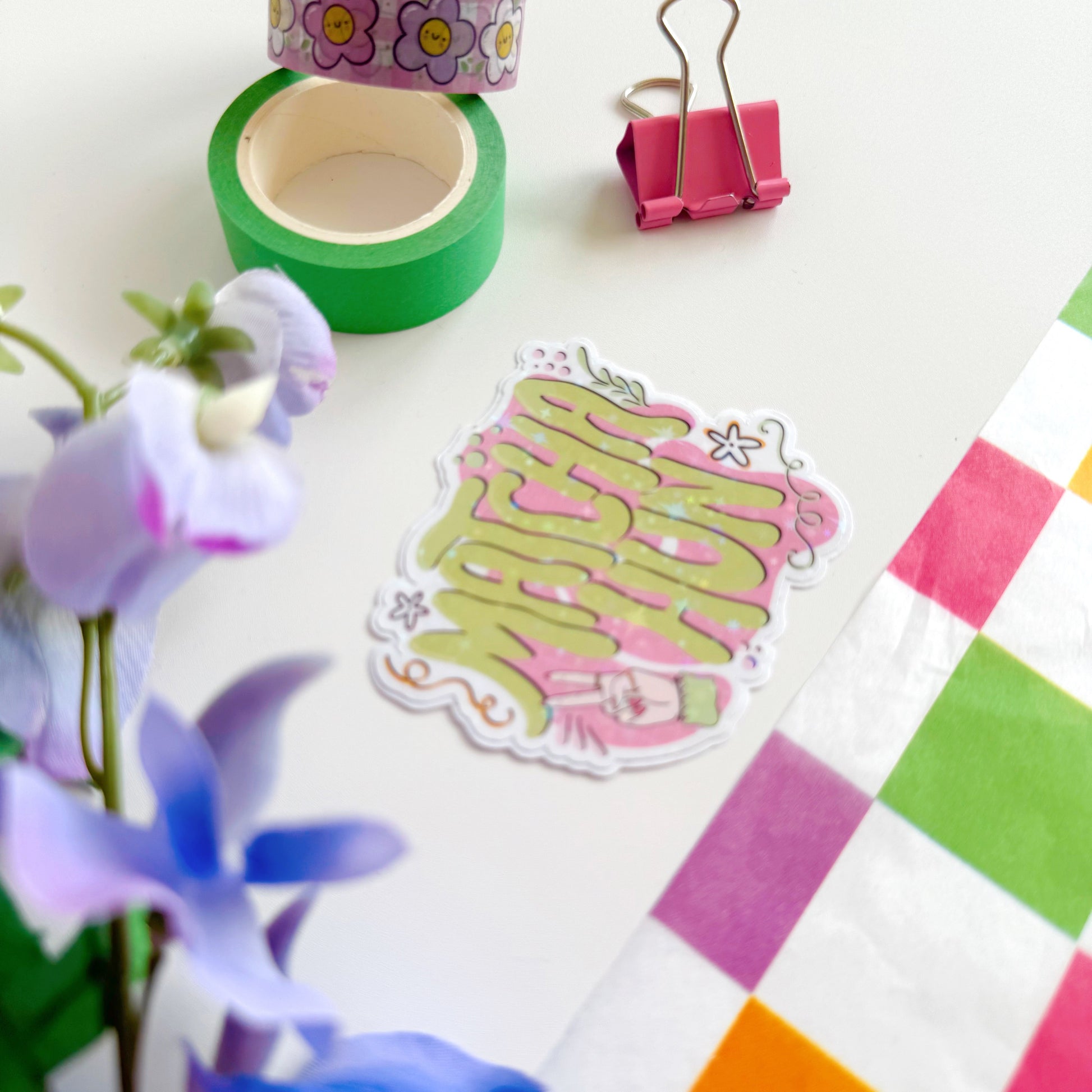 A holographic sticker with text saying 'Matcha Hun' in funky bubble writing and other hand illustrated elements around the text. The colours are pink and green.