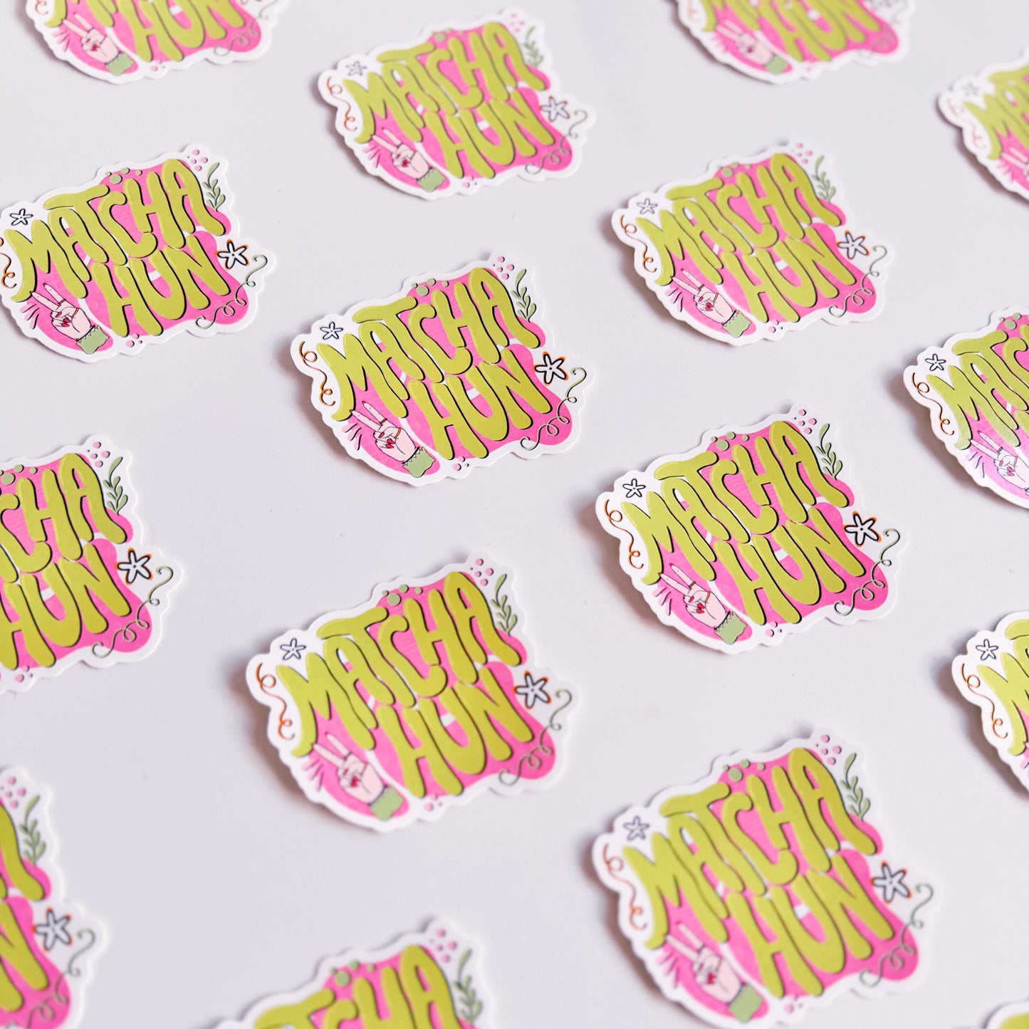 Several vinyl stickers on a white background. The vinyl sticker is pink and green and says the words 'Matcha Hun' in fun bubble writing. 