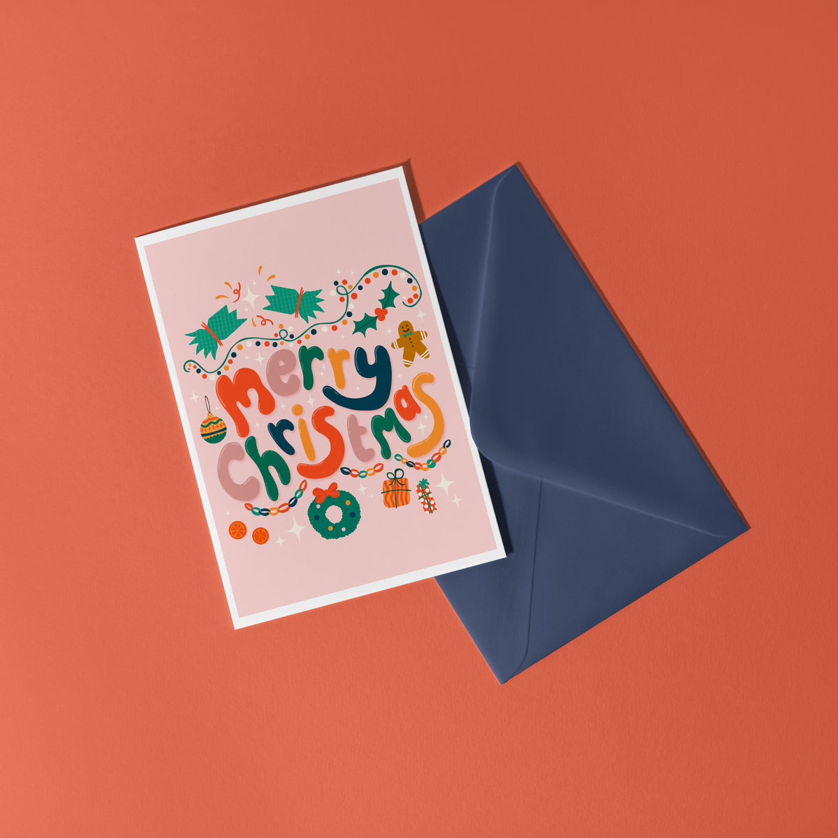 A dusky pink Christmas greeting card with colourful retro-style lettering and festive illustrations, styled on a light surface with silver tinsel, miniature wrapped gifts, and holiday decorations.