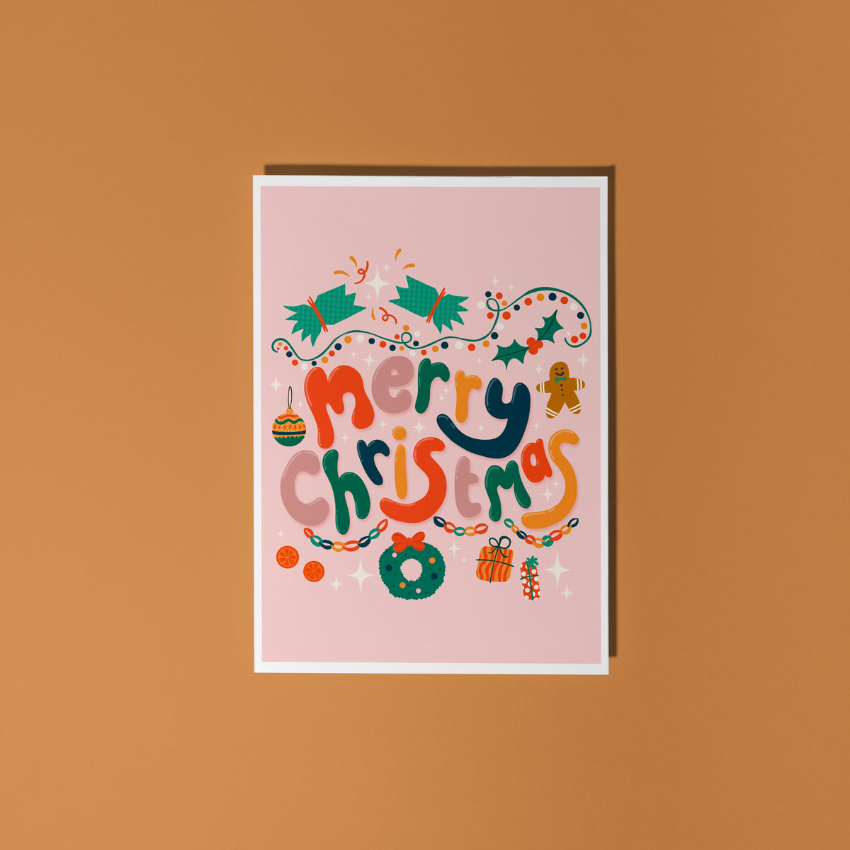 A dusky pink Christmas greeting card with colourful retro-style lettering and festive illustrations, styled on a light surface with silver tinsel, miniature wrapped gifts, and holiday decorations.