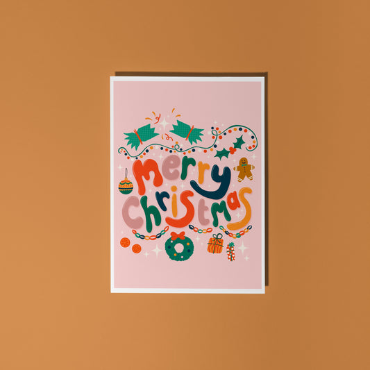 A dusky pink Christmas greeting card with colourful retro-style lettering and festive illustrations, styled on a light surface with silver tinsel, miniature wrapped gifts, and holiday decorations.
