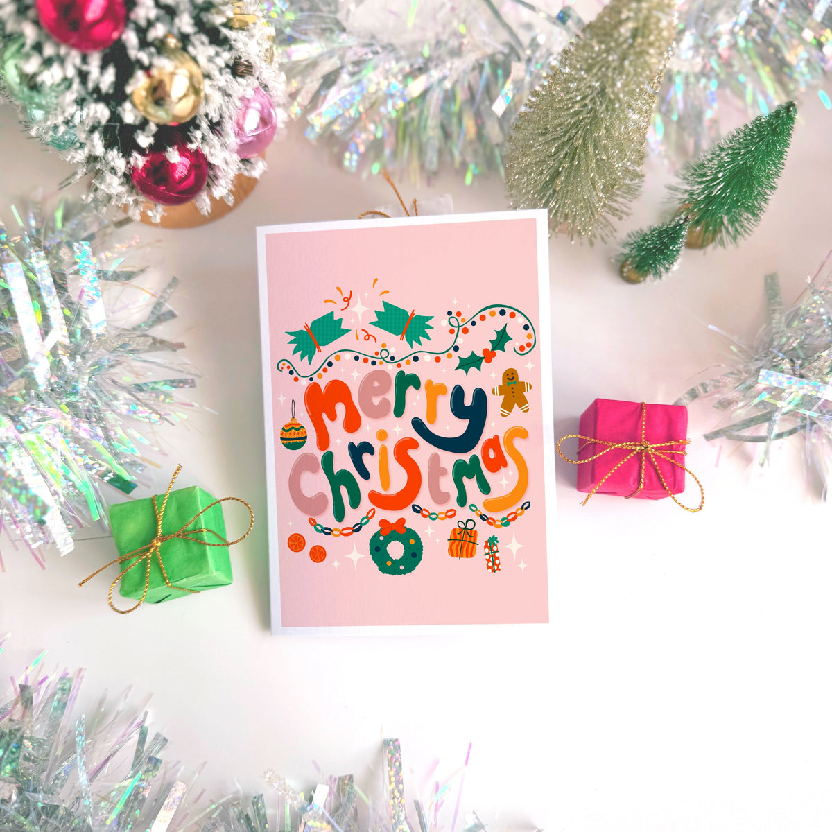 A dusky pink Christmas greeting card with colourful retro-style lettering and festive illustrations, styled on a light surface with silver tinsel, miniature wrapped gifts, and holiday decorations.