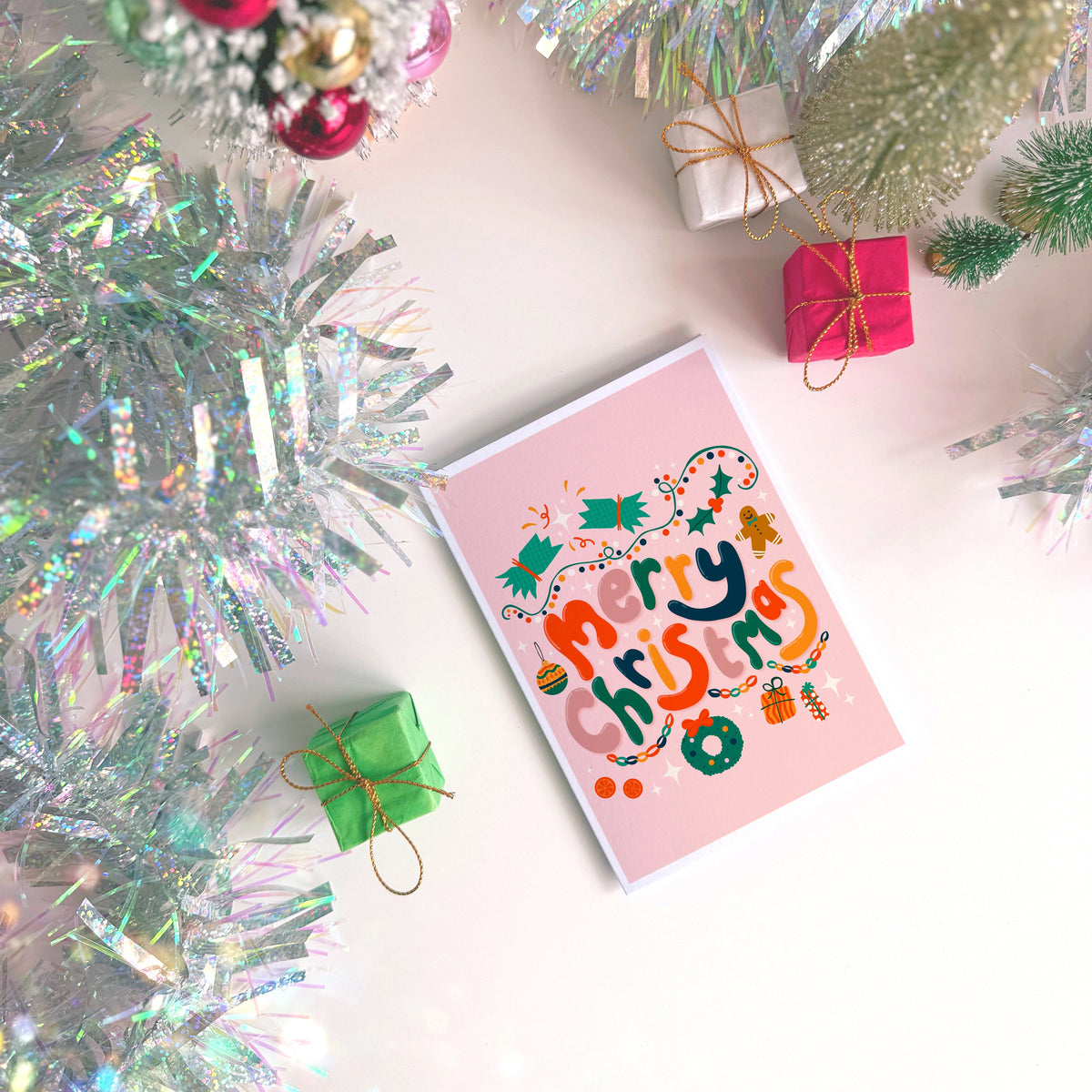 A dusky pink Christmas greeting card with colourful retro-style lettering and festive illustrations, styled on a light surface with silver tinsel, miniature wrapped gifts, and holiday decorations.