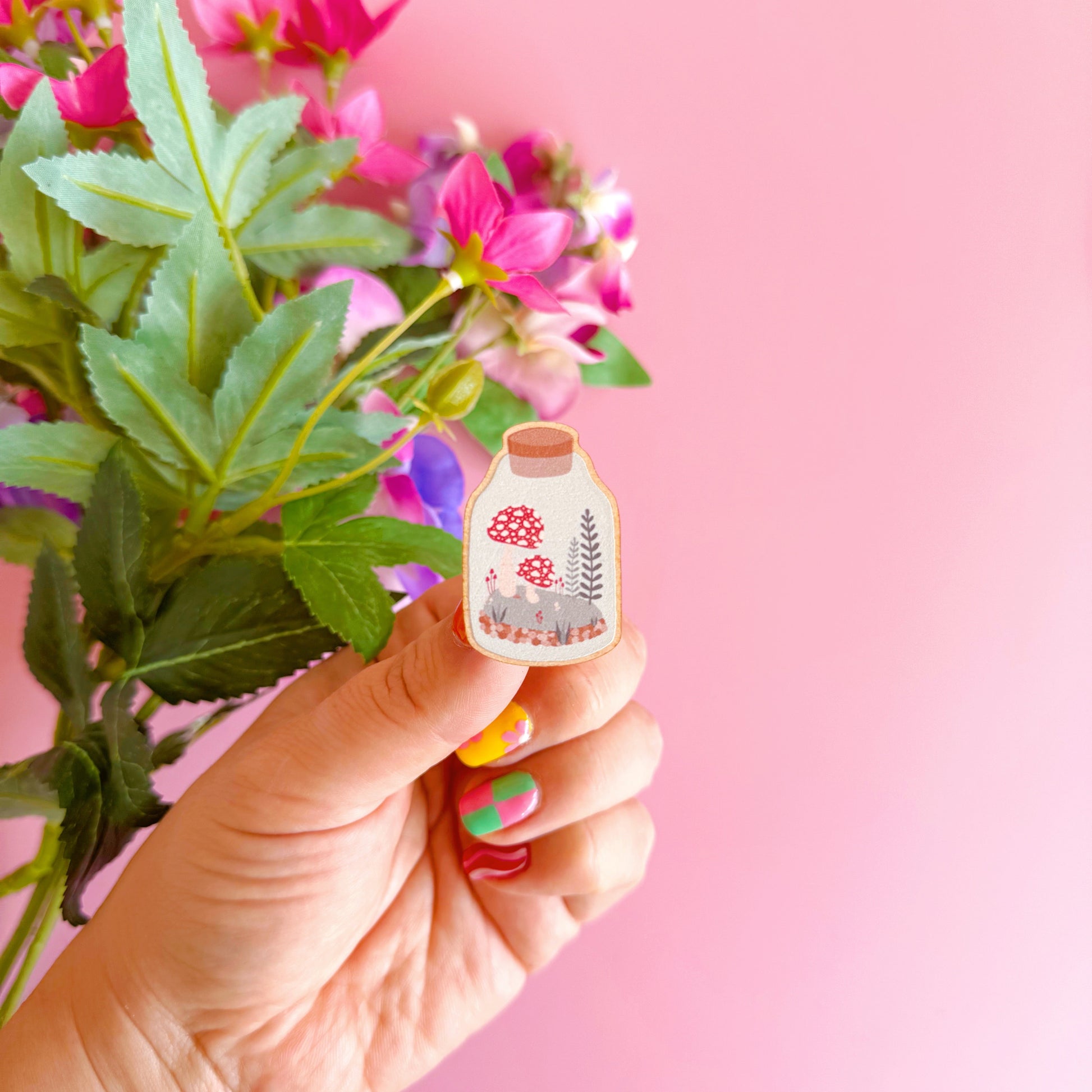 A wooden pin badge featuring an illustration of a terrarium filled with mushrooms and woodland elements.