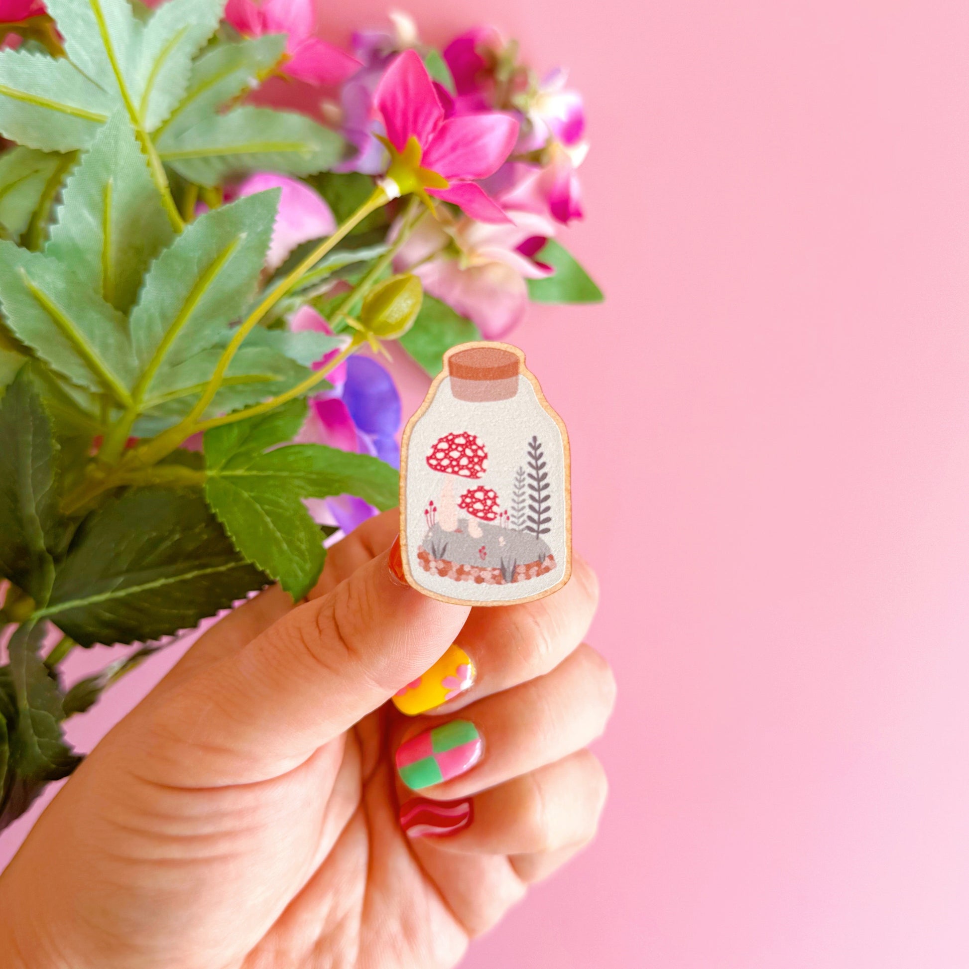 A terrarium jar illustration, with a mini woodland scene with mushrooms and ferns inside, on an eco friendly wooden pin badge. 