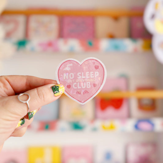 A pink holographic sticker, in the shape of a heart, with the words 'No sleep (but surviving) club' in the centre. Around the text are little doodles, like tired eyes, hearts and a moon behind clouds.