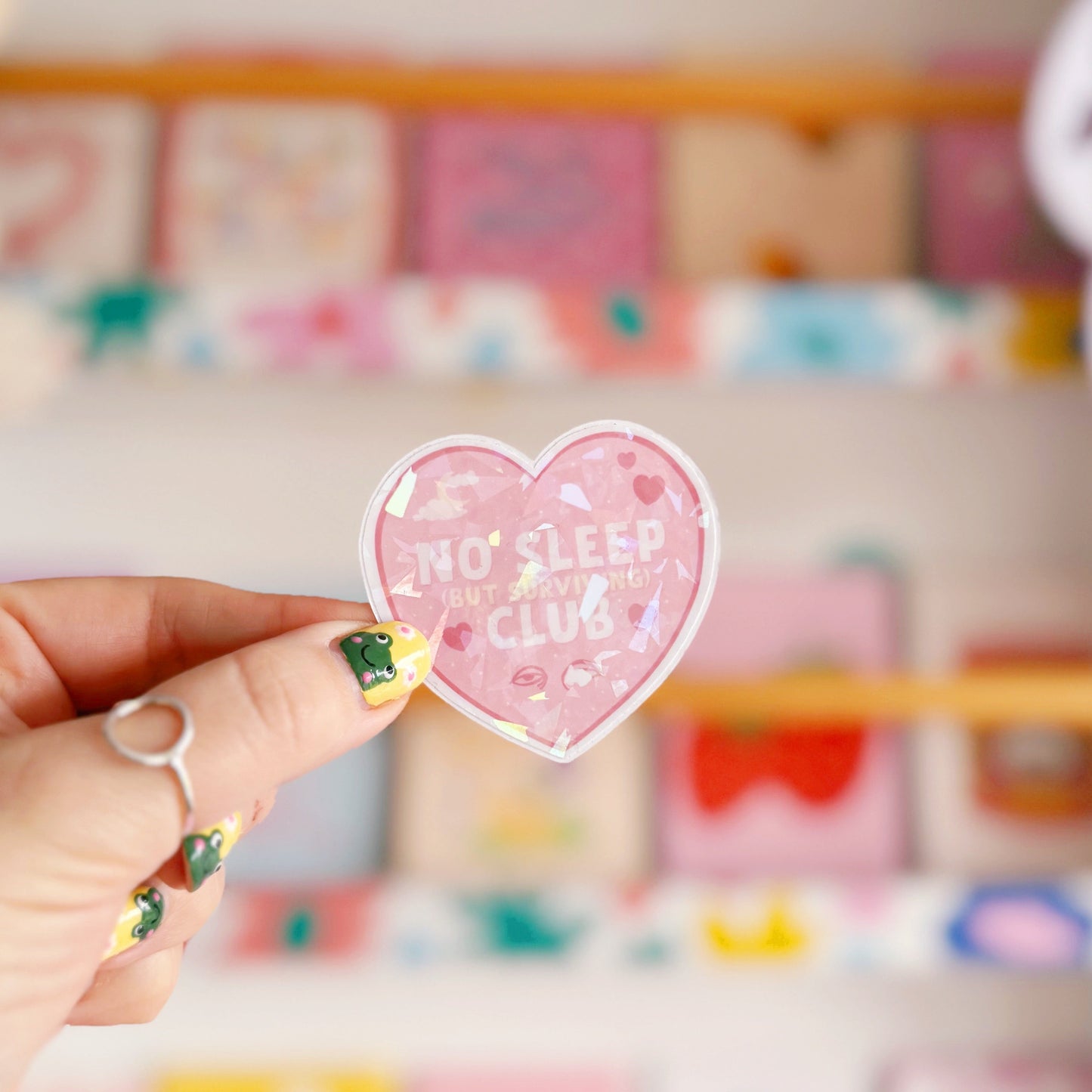 A pink holographic sticker, in the shape of a heart, with the words 'No sleep (but surviving) club' in the centre. Around the text are little doodles, like tired eyes, hearts and a moon behind clouds.