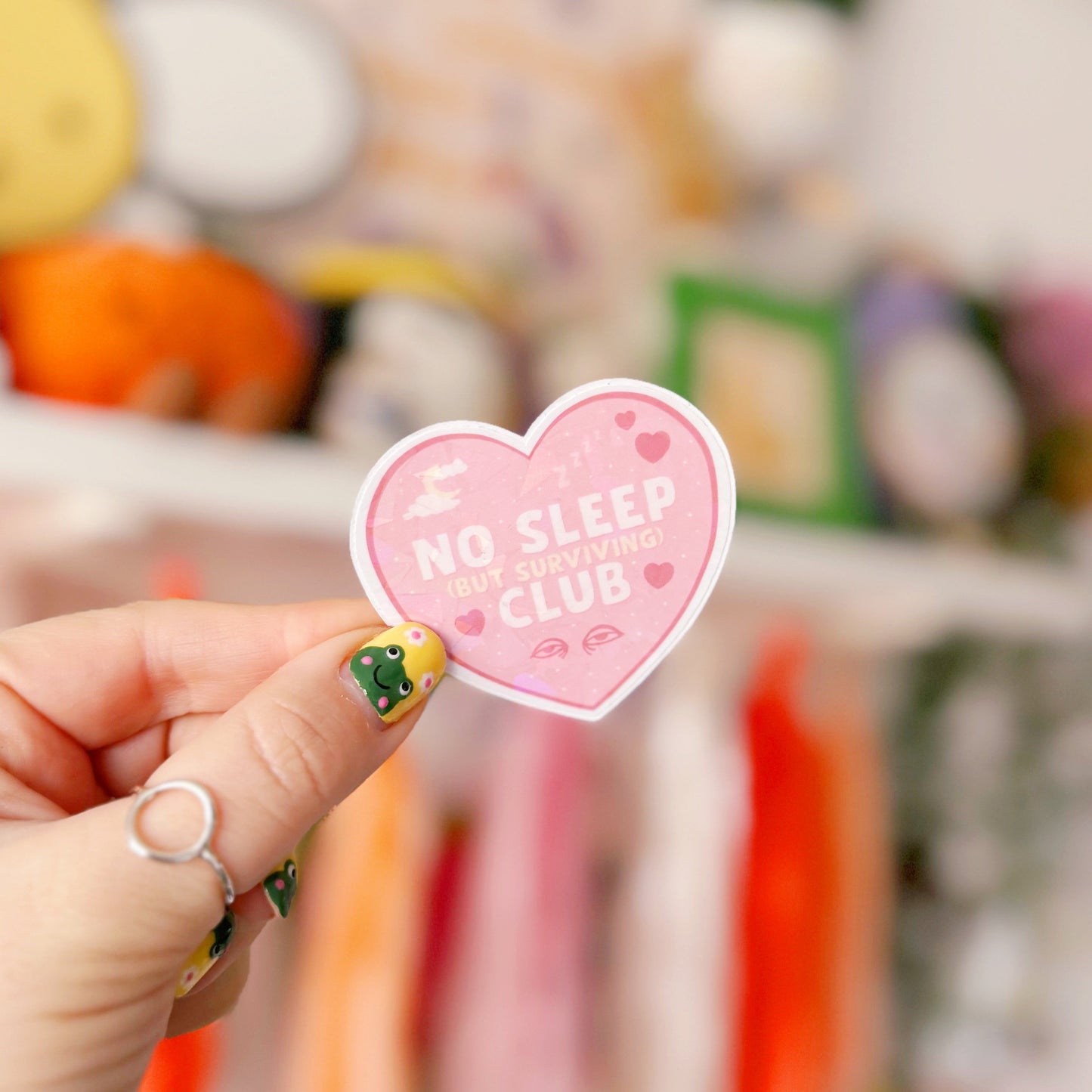 A pink holographic sticker, in the shape of a heart, with the words 'No sleep (but surviving) club' in the centre. Around the text are little doodles, like tired eyes, hearts and a moon behind clouds.