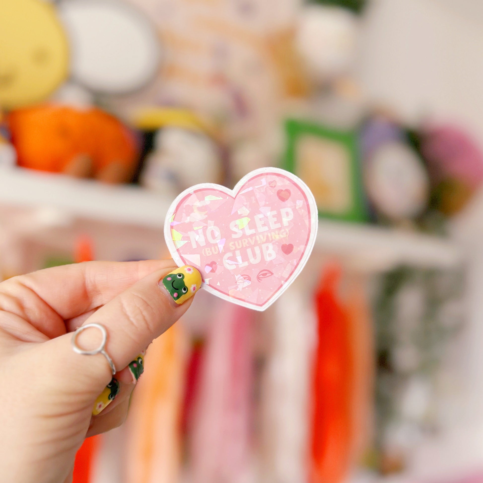 A pink holographic sticker, in the shape of a heart, with the words 'No sleep (but surviving) club' in the centre. Around the text are little doodles, like tired eyes, hearts and a moon behind clouds.
