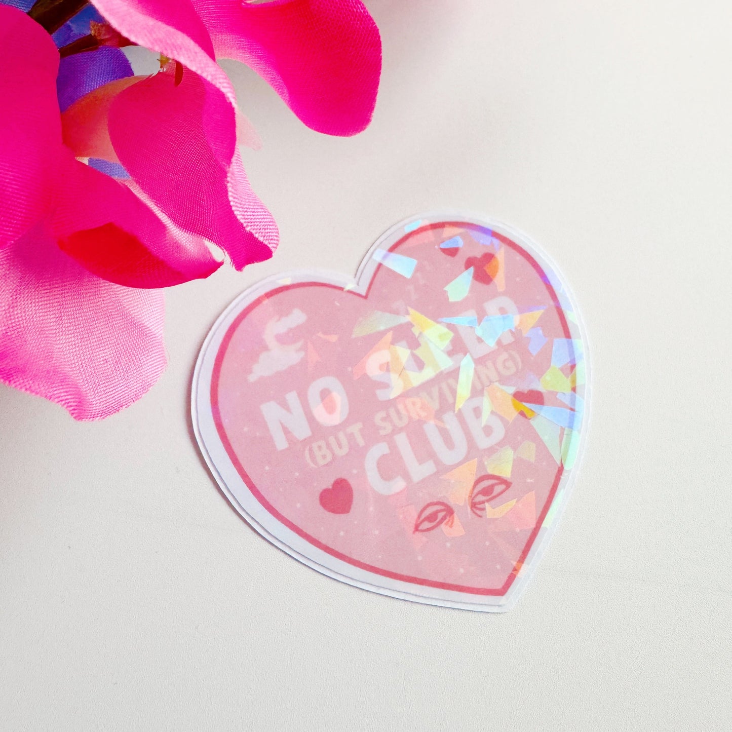 A holographic sticker, a pink heart with the words 'no sleep but surviving club' in. The sticker has an easy peel lip.