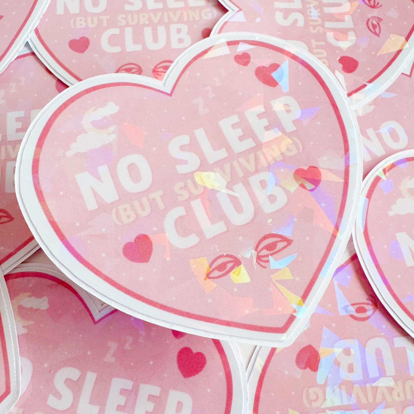 A holographic sticker, a pink heart with the words 'no sleep but surviving club' in. The sticker has an easy peel lip.