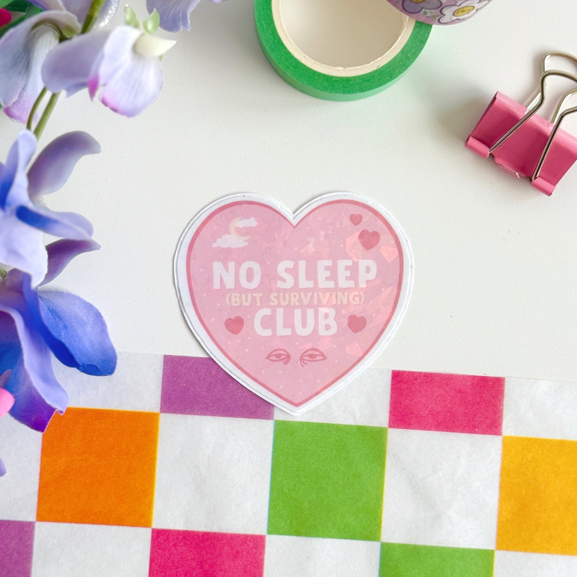A pink holographic sticker, in the shape of a heart, with the words 'No sleep (but surviving) club' in the centre. Around the text are little doodles, like tired eyes, hearts and a moon behind clouds.