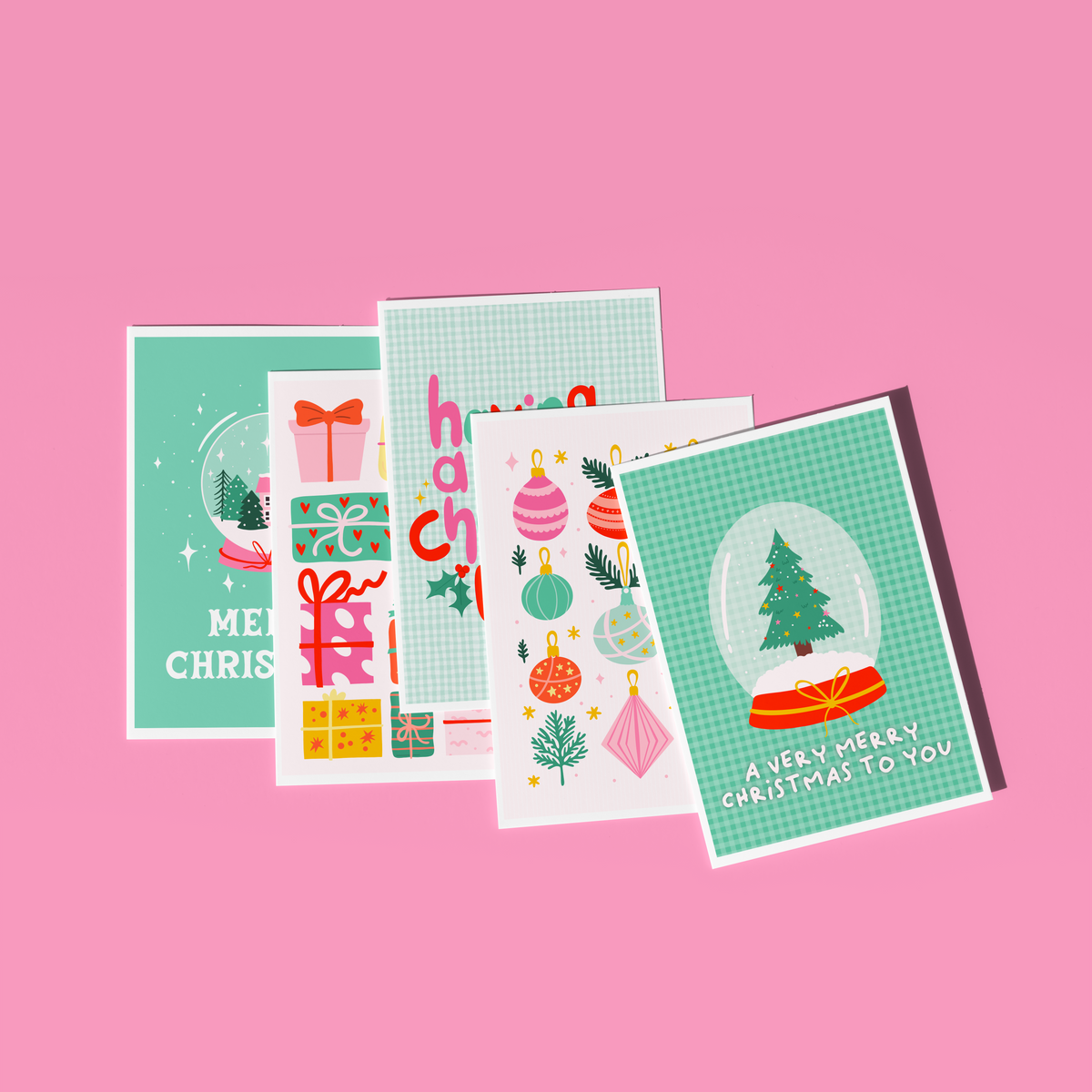 Four Christmas greeting cards arranged in a fan-like overlap on a solid pink background. The cards feature festive illustrations including ornaments, gifts, a snow globe with a Christmas tree, and holiday text such as 'Merry Christmas' and 'A Very Merry Christmas to You.