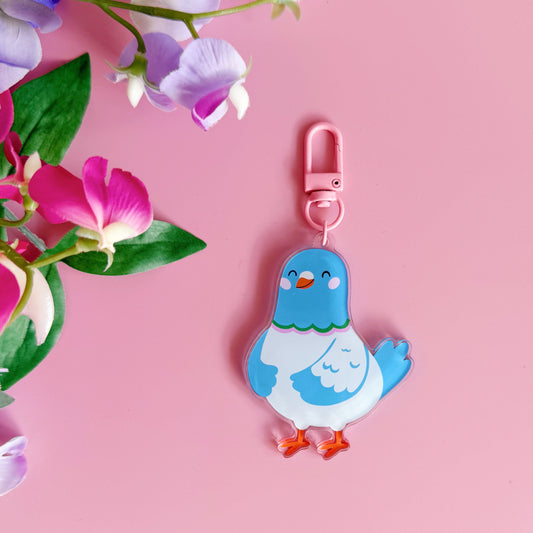 Blue cartoon pigeon keychain with a pink clip on a pink background with flowers.