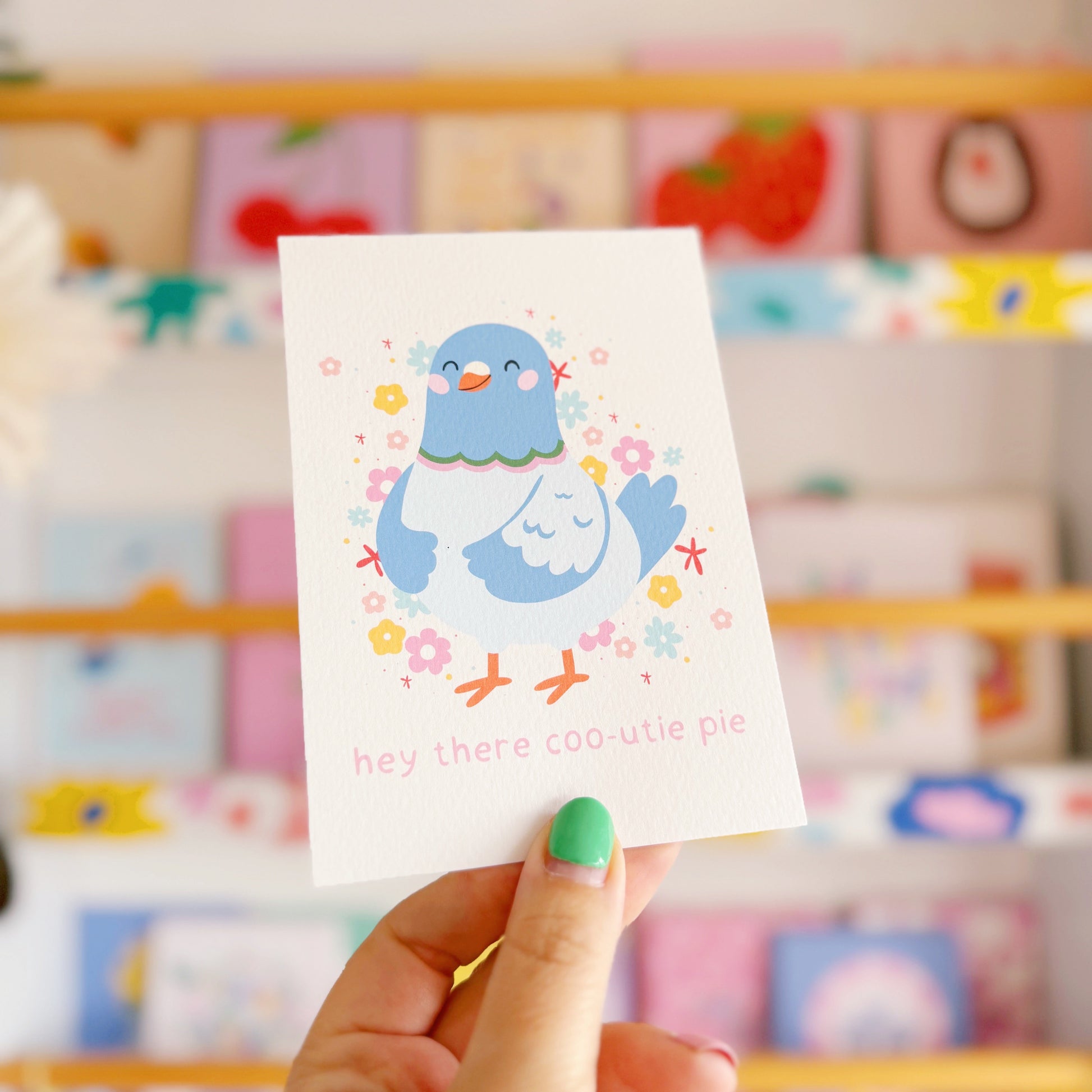 Card with a cartoon pigeon holding flowers, held by a hand against a colourful background.