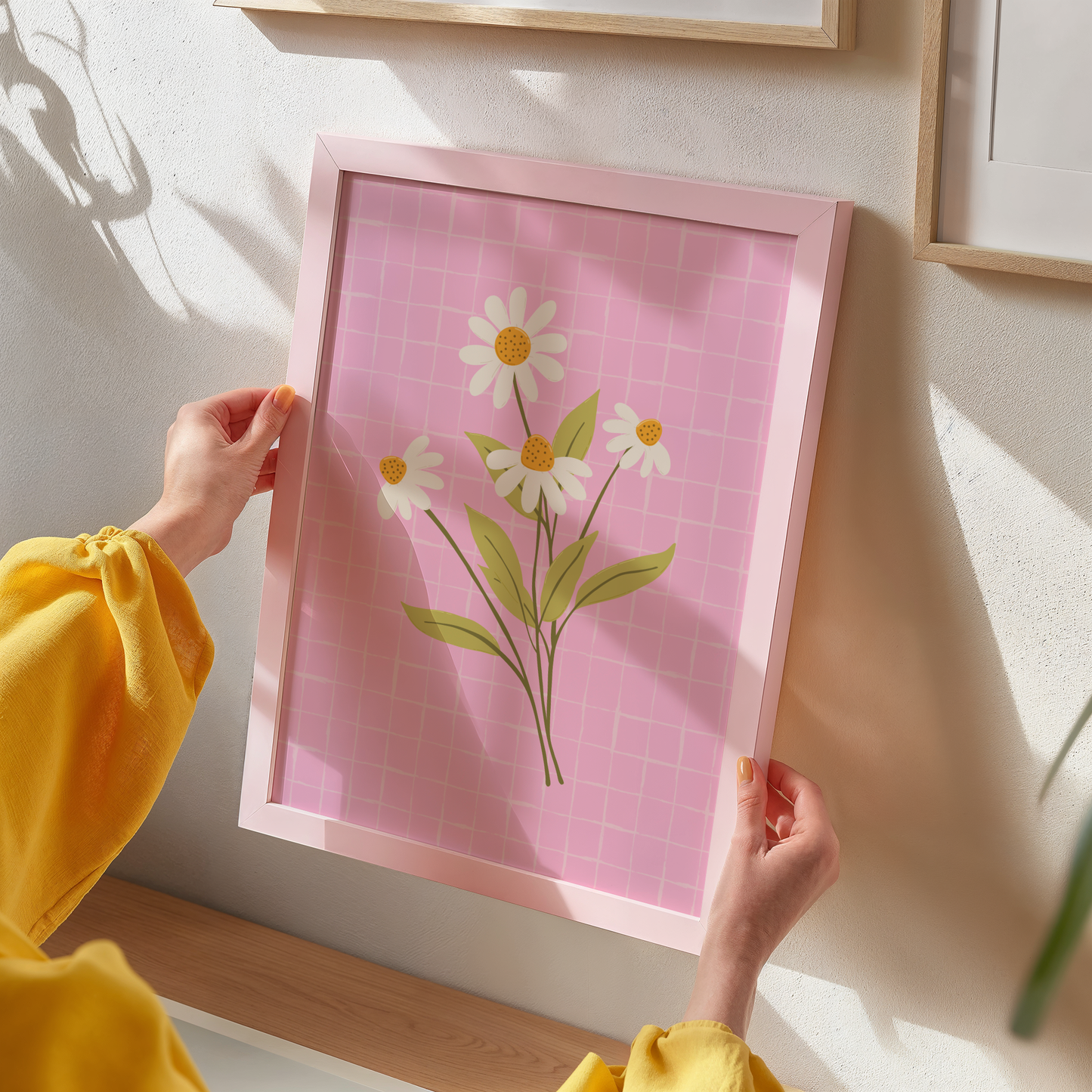 Person holding a framed print of daisies on a pink grid background