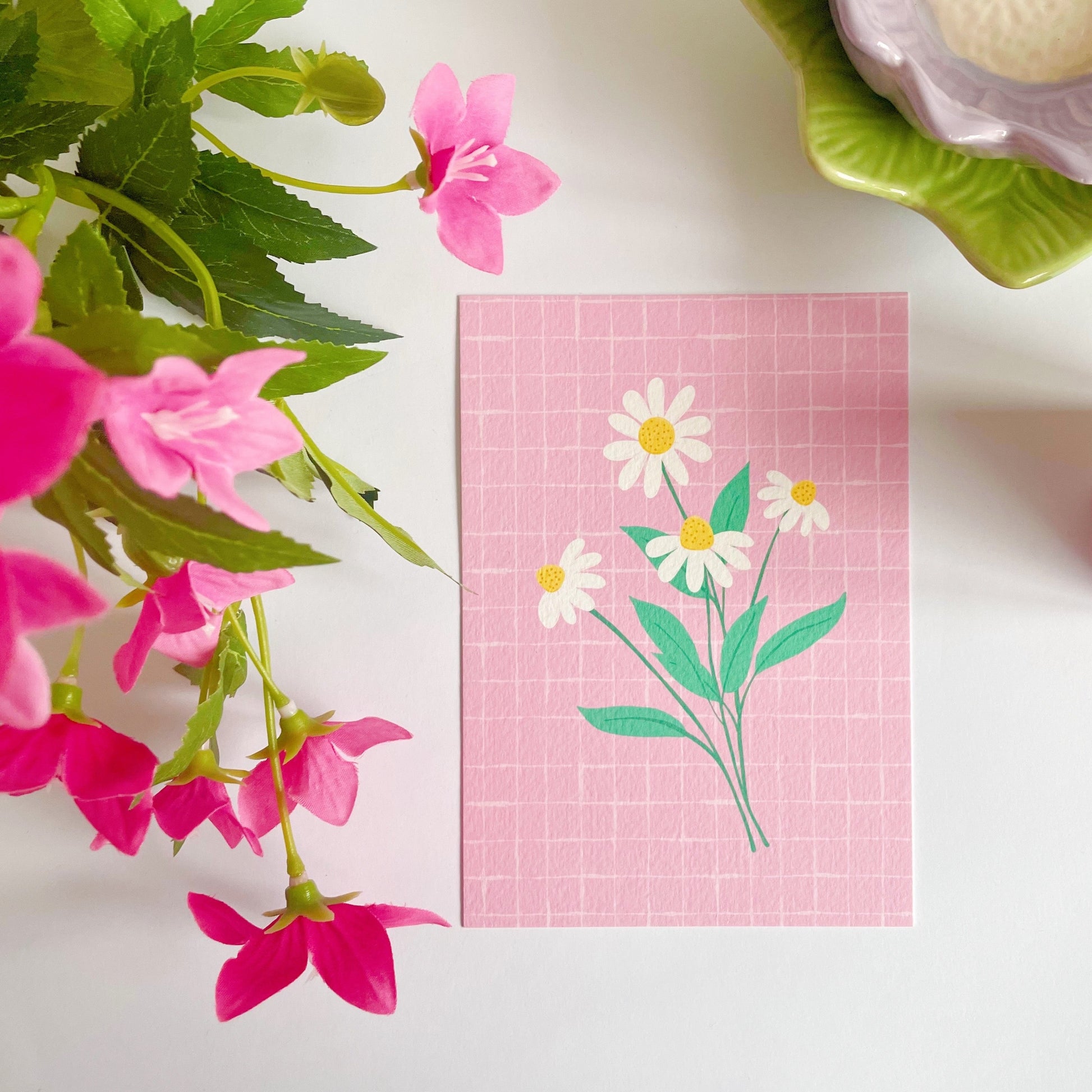 A pink textured postcard with an illustration of four daisies with bright green stems.