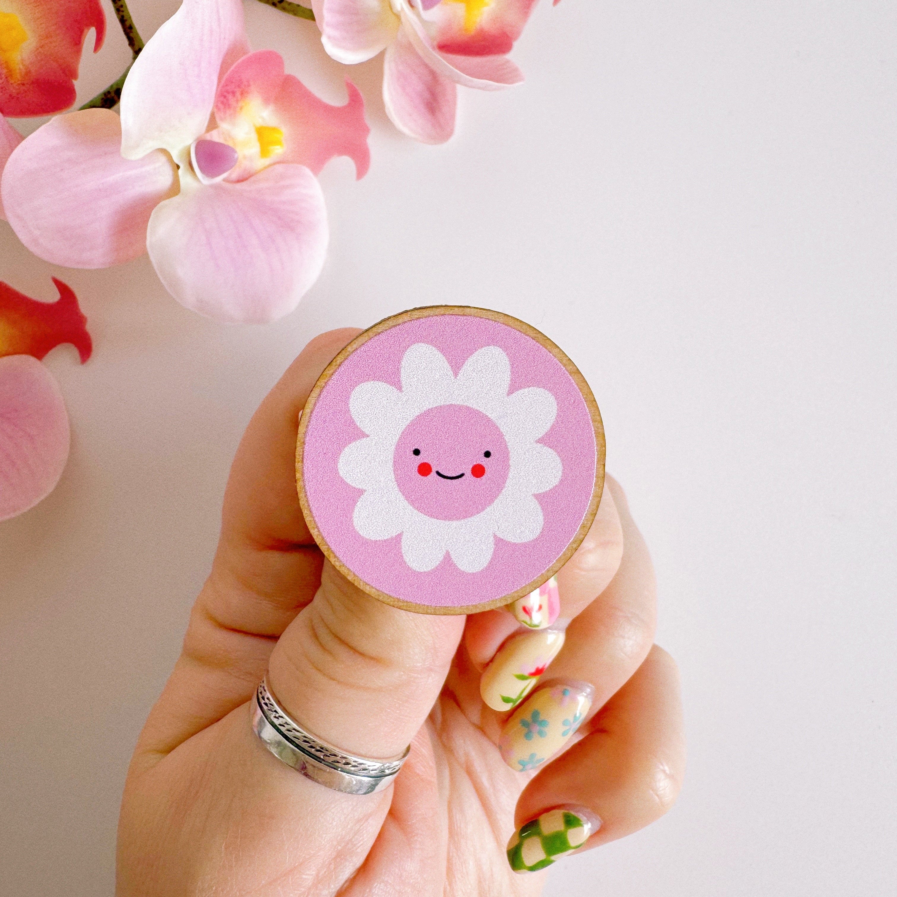 A pink pin badge, of a pink circle with a white daisy in the centre. the daisy is smiling and has rosy red cheeks. the backing card of the pin badge is yellow checkerboard patterned and has branding on.