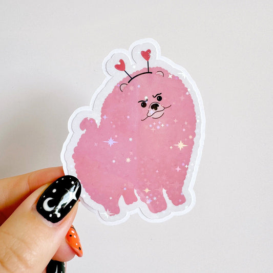 A holographic sticker, of a pink grumpy looking dog with a heart shaped headband on. The sticker has an easy peel lip.