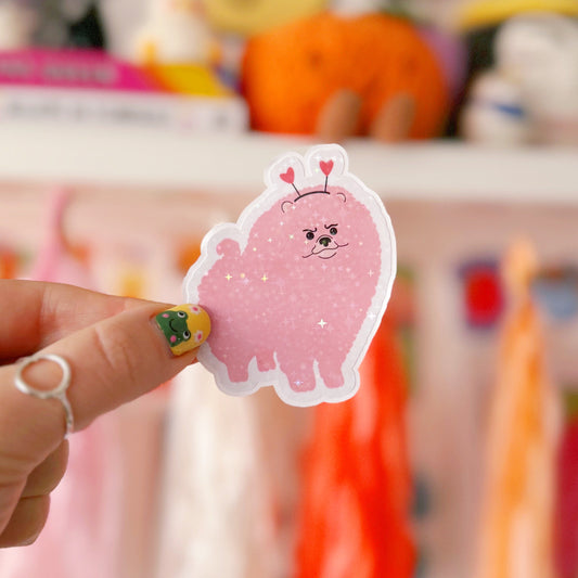 A holographic sticker, of a pink grumpy looking dog with a heart shaped headband on. The sticker has an easy peel lip.
