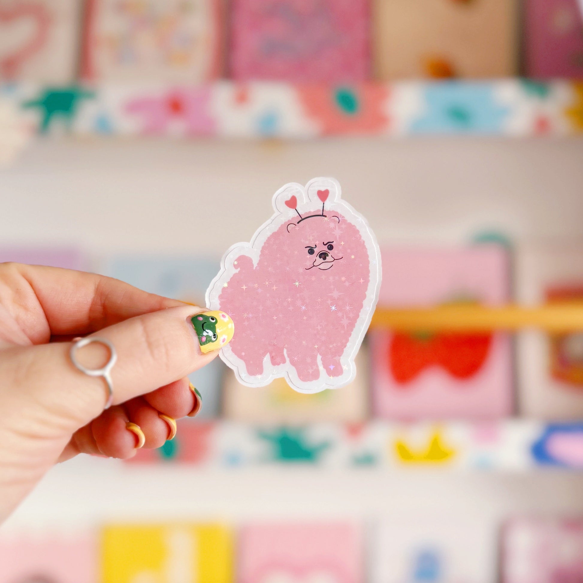 A holographic sticker, of a pink grumpy looking dog with a heart shaped headband on. The sticker has an easy peel lip.