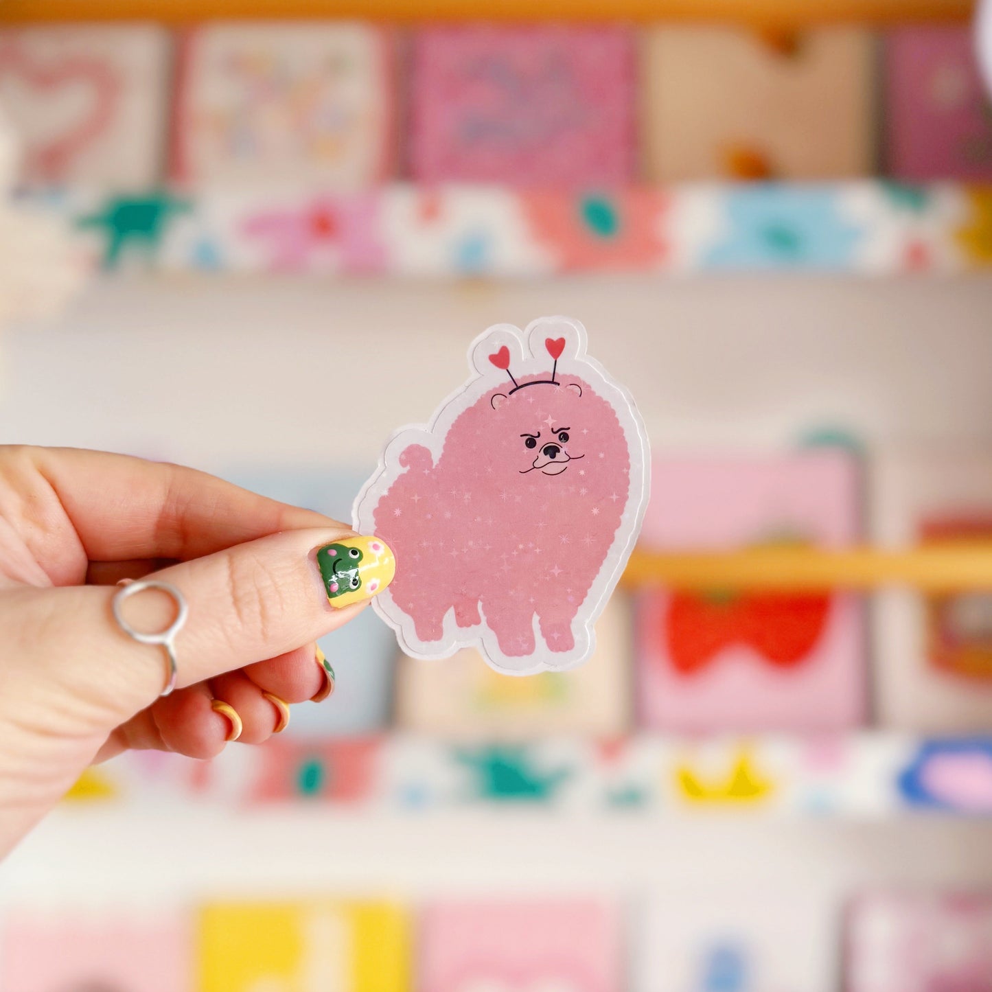 A holographic sticker, of a pink grumpy looking dog with a heart shaped headband on. The sticker has an easy peel lip.