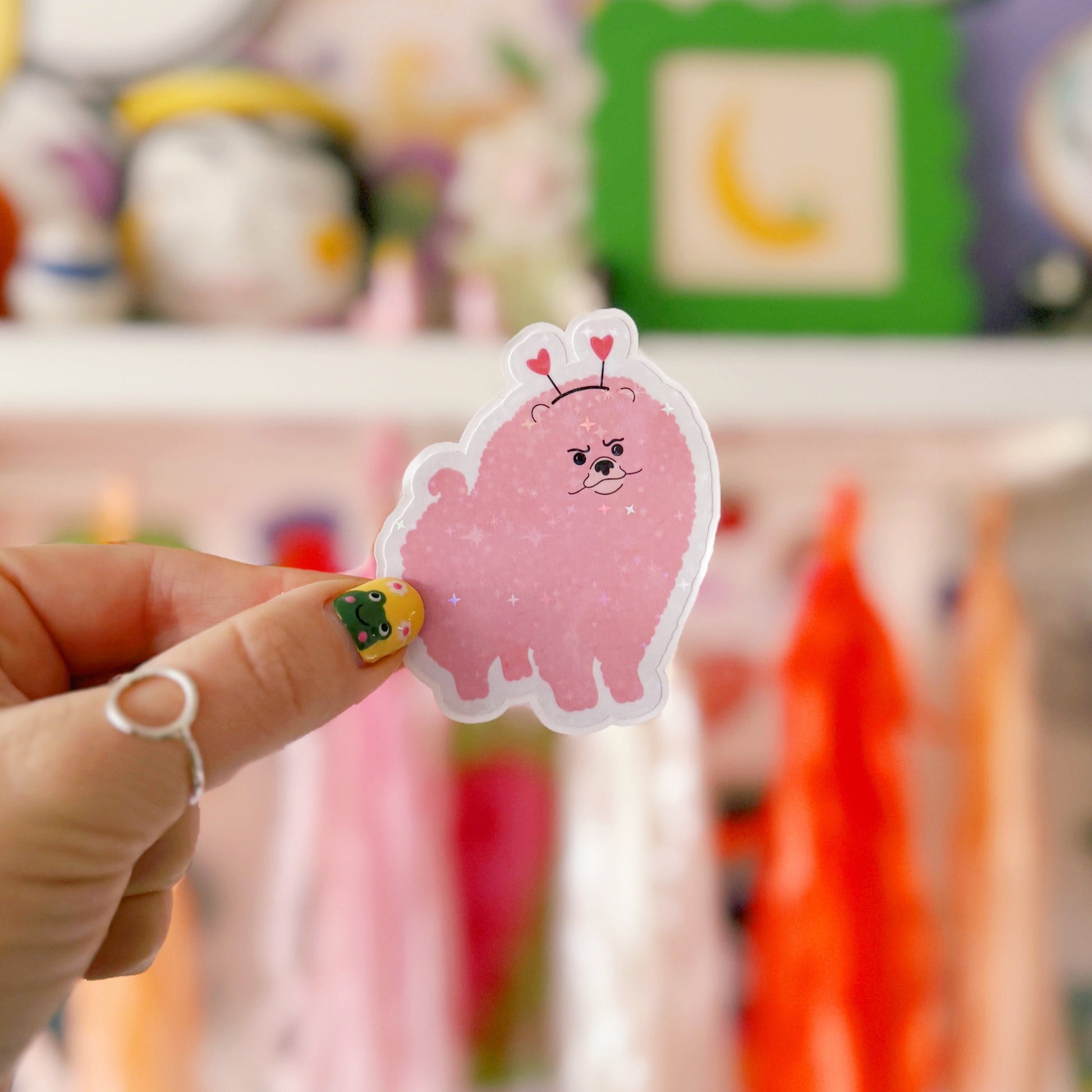 A holographic sticker, of a pink grumpy looking dog with a heart shaped headband on. The sticker has an easy peel lip.