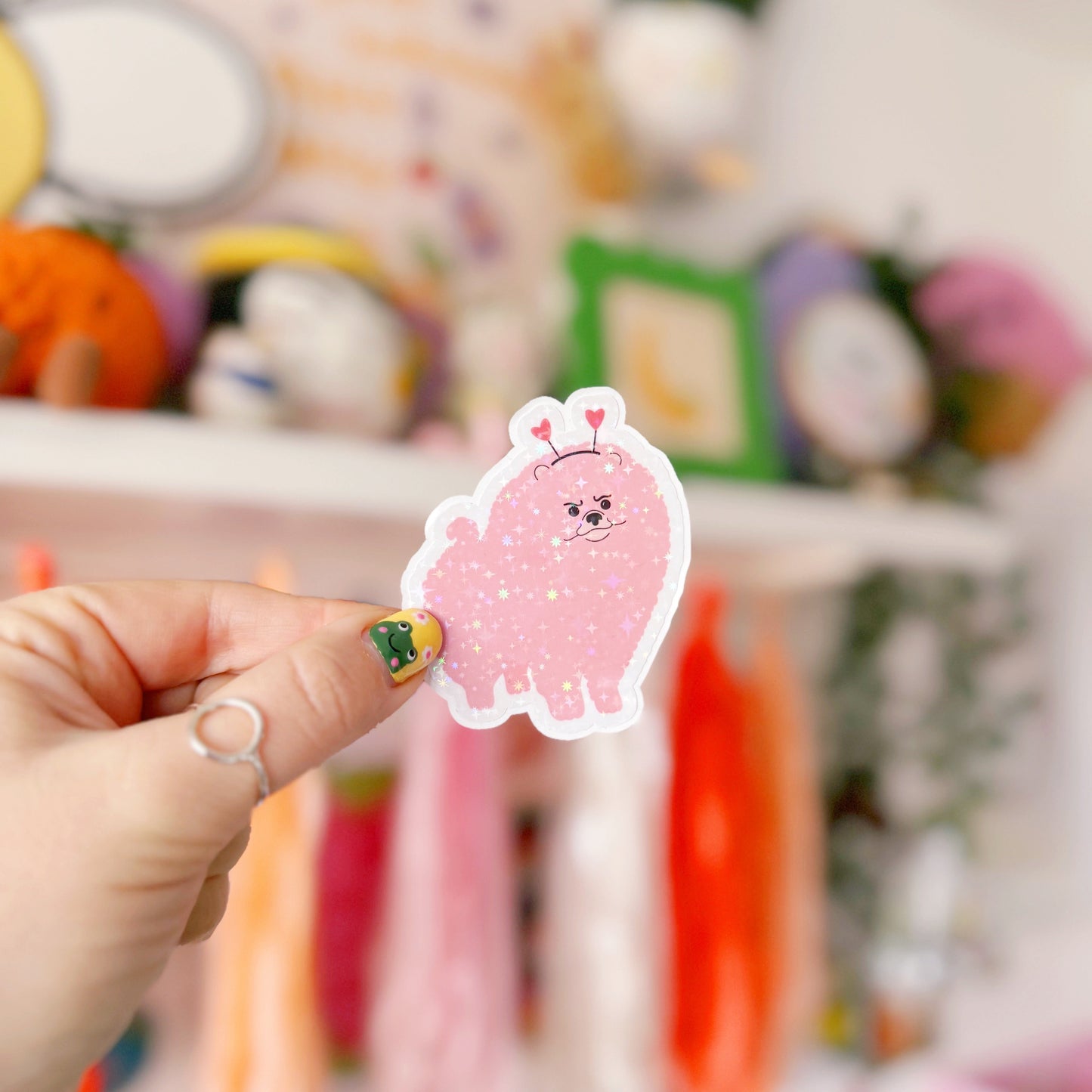 A holographic sticker, of a pink grumpy looking dog with a heart shaped headband on. The sticker has an easy peel lip.