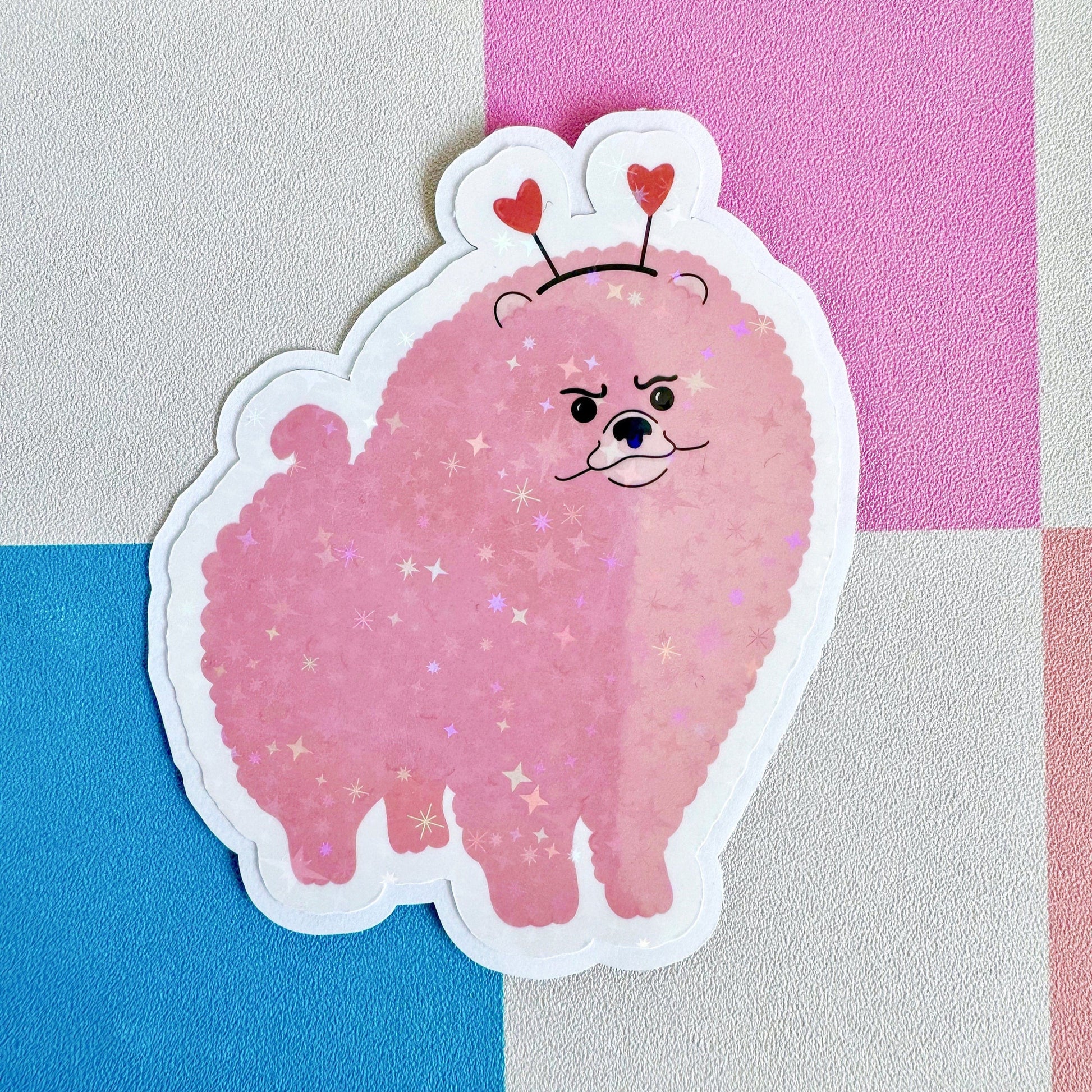 A holographic sticker, of a pink grumpy looking dog with a heart shaped headband on. The sticker has an easy peel lip.