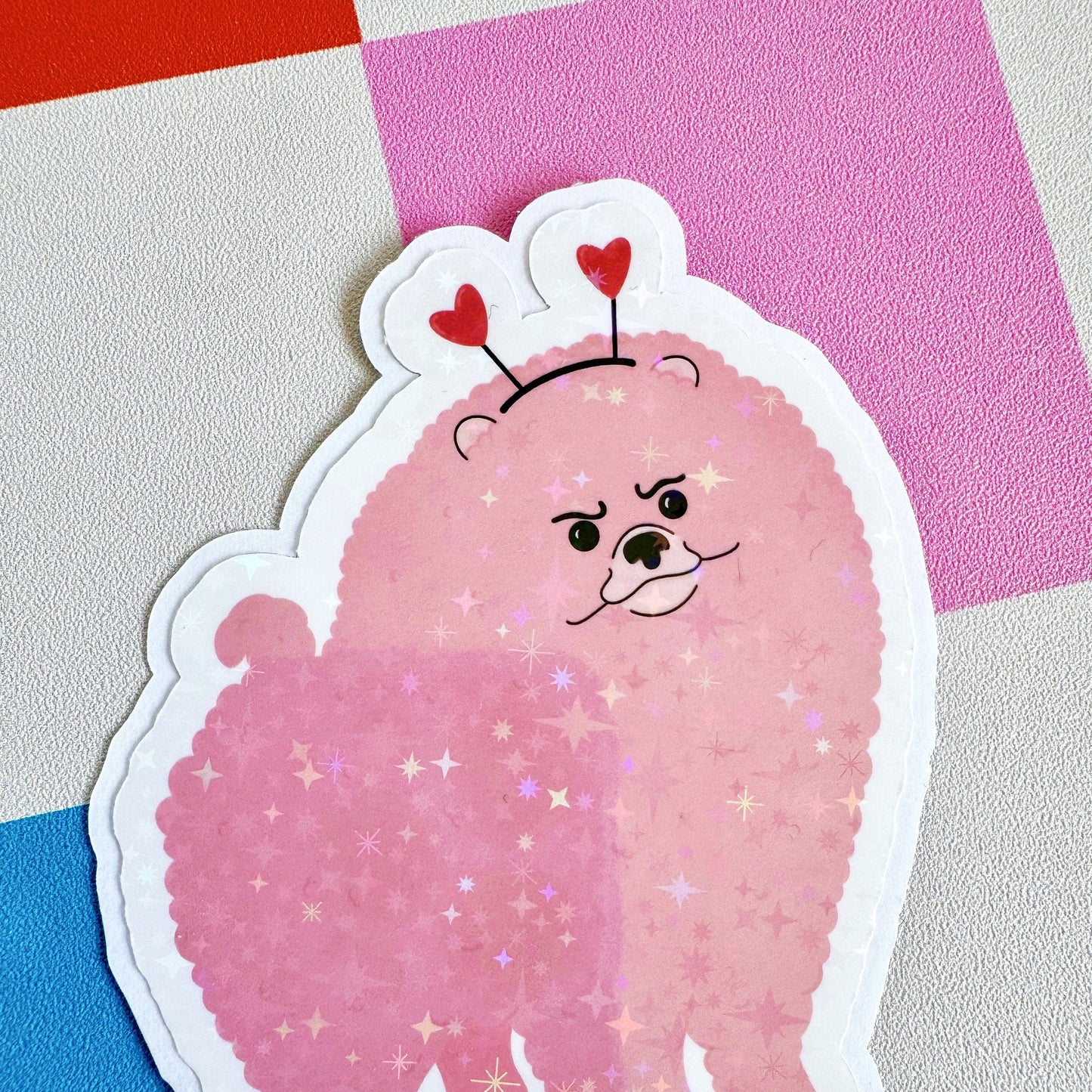 A holographic sticker, of a pink grumpy looking dog with a heart shaped headband on. The sticker has an easy peel lip.