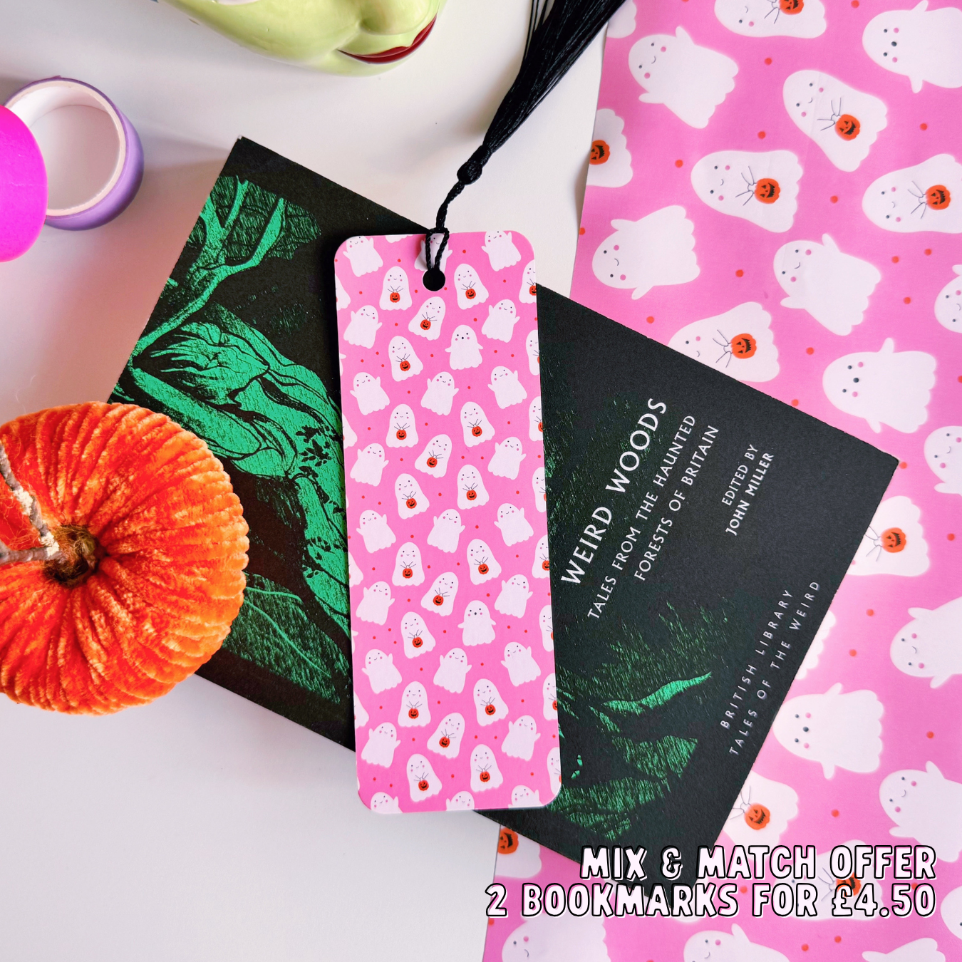 A styled flat-lay featuring a pink paper bookmark with a black tassel, printed with a repeating pattern of white ghosts with pumpkin trick or treat baskets. 