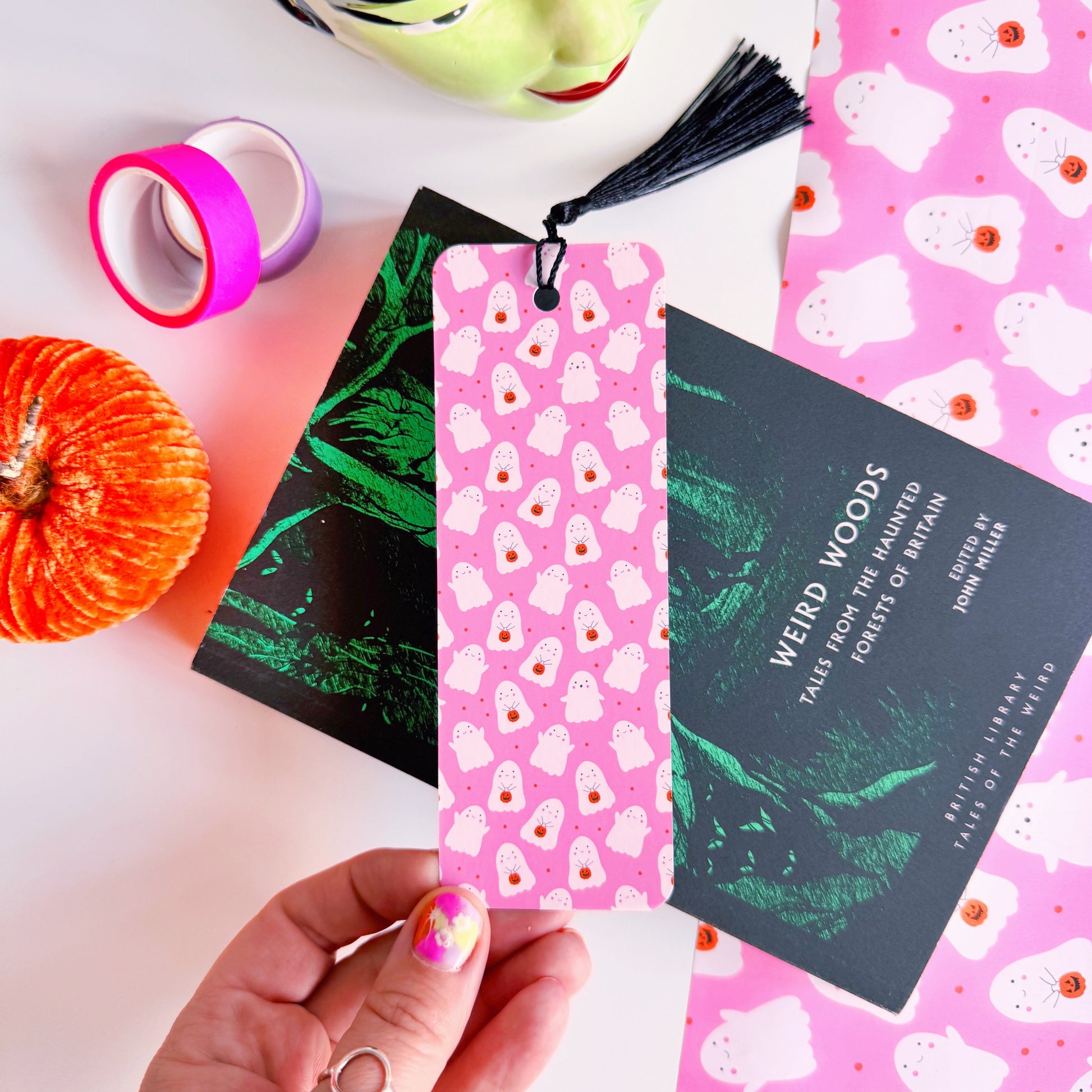 A styled flat-lay featuring a pink paper bookmark with a black tassel, printed with a repeating pattern of white ghosts with pumpkin trick or treat baskets. 