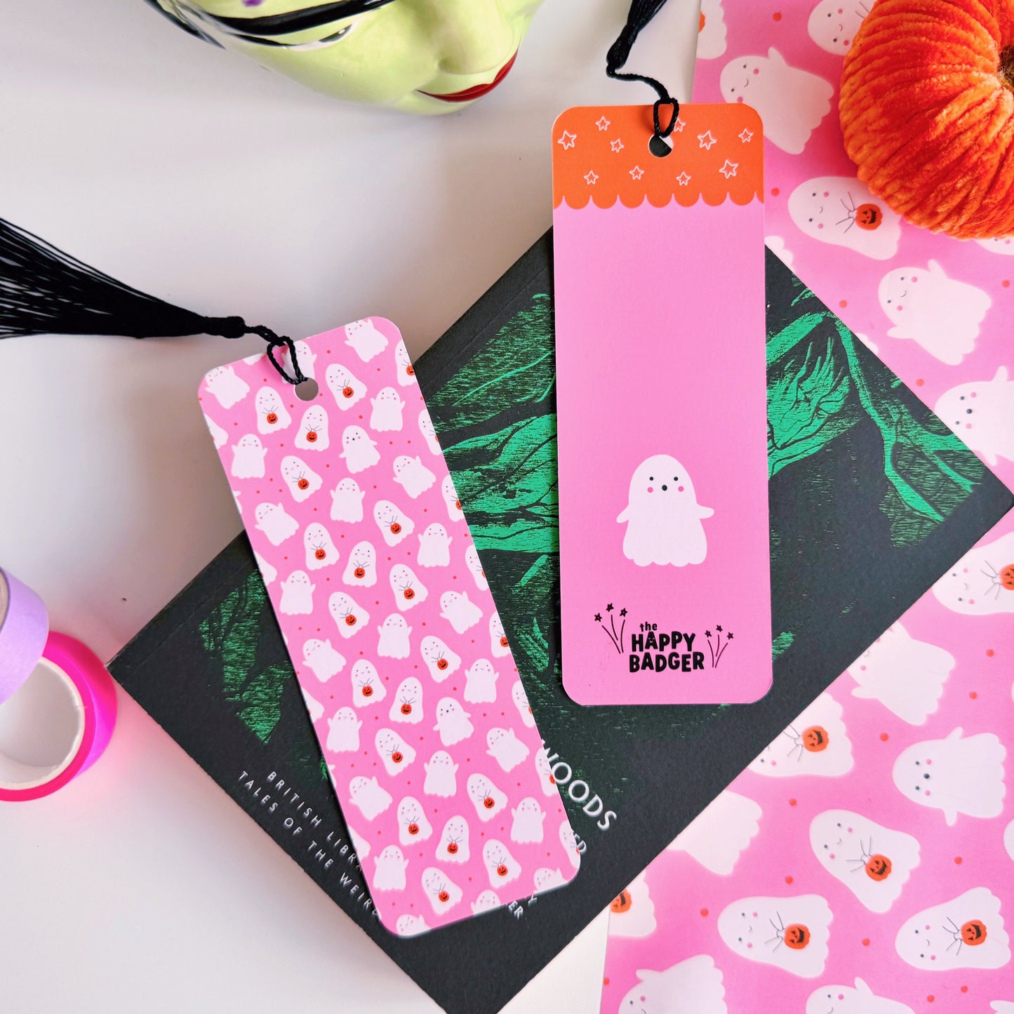 A styled flat-lay featuring a pink paper bookmark with a black tassel, printed with a repeating pattern of white ghosts with pumpkin trick or treat baskets. 