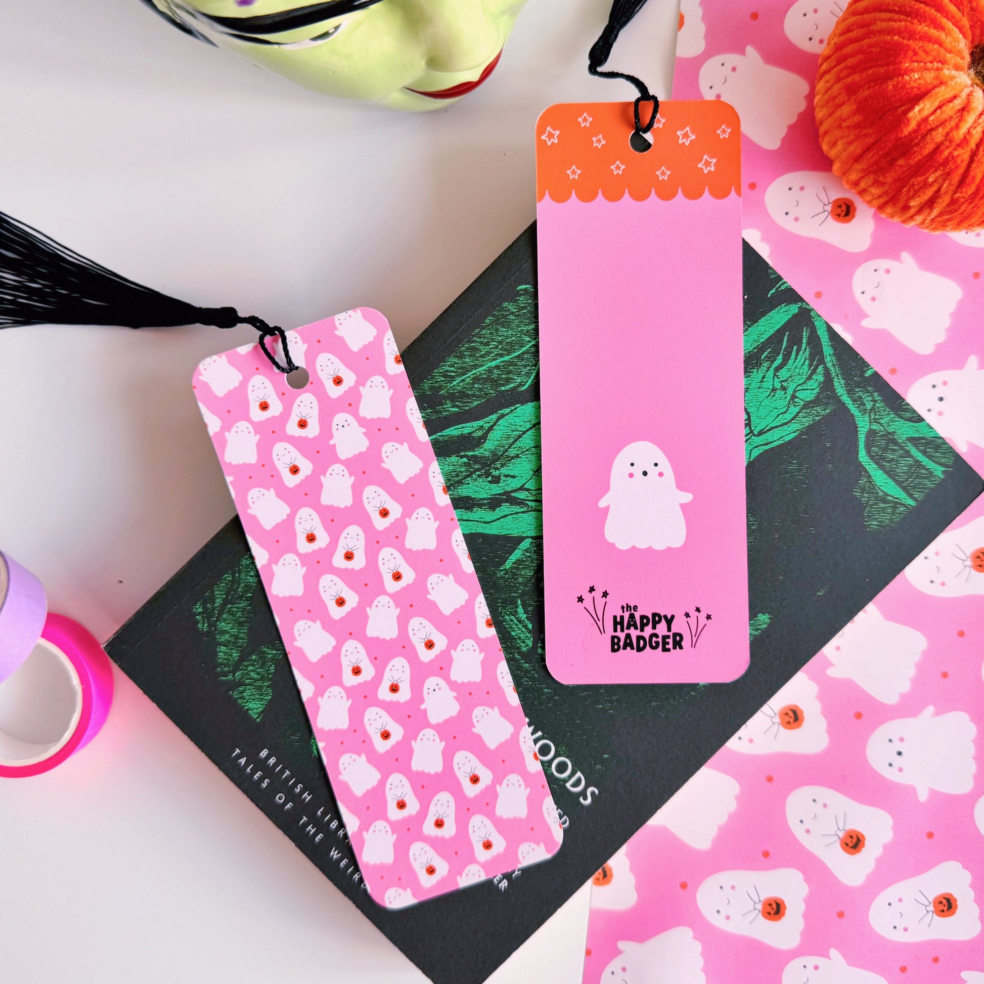 A styled flat-lay featuring a pink paper bookmark with a black tassel, printed with a repeating pattern of white ghosts with pumpkin trick or treat baskets. 