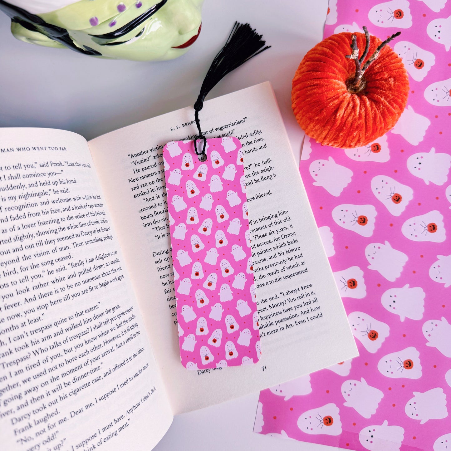 A styled flat-lay featuring a pink paper bookmark with a black tassel, printed with a repeating pattern of white ghosts with pumpkin trick or treat baskets. 