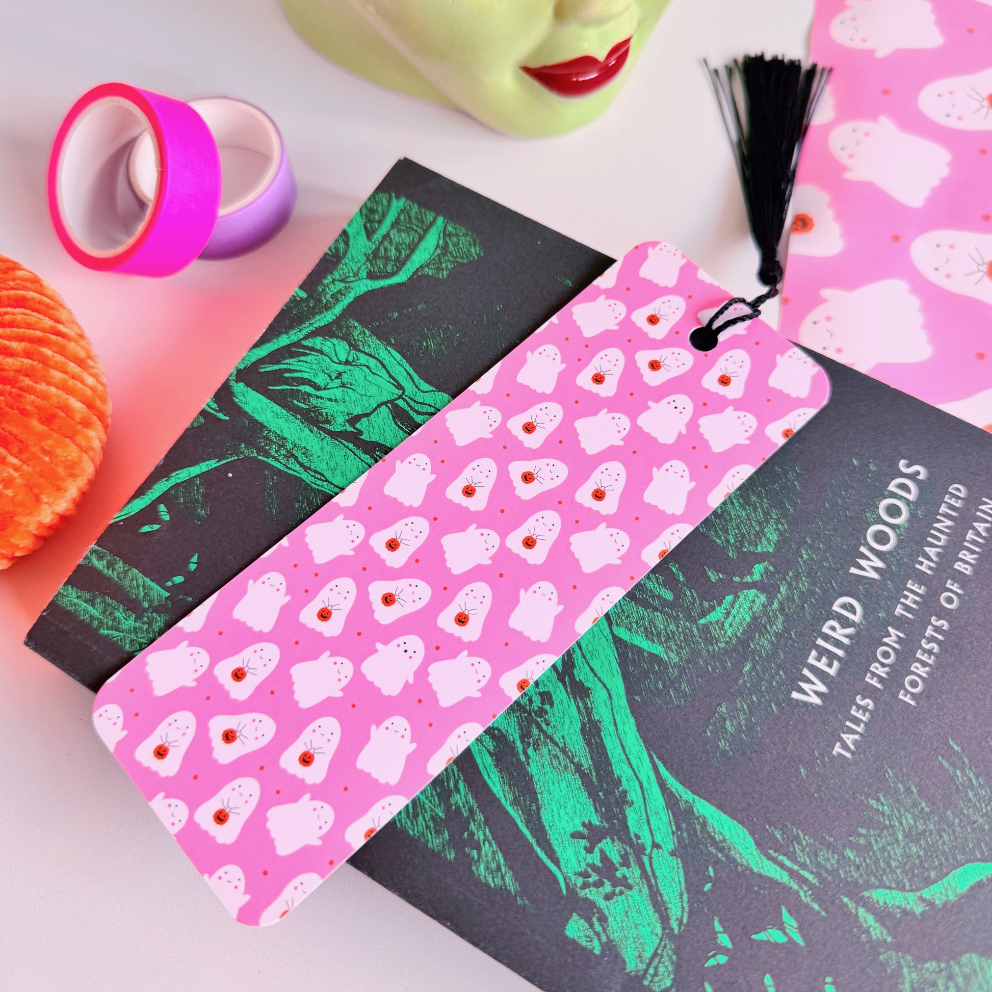 A styled flat-lay featuring a pink paper bookmark with a black tassel, printed with a repeating pattern of white ghosts with pumpkin trick or treat baskets. 