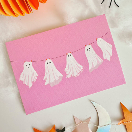 A halloween themed postcard of 5 ghosts hanging on a washing line, on a pink background.