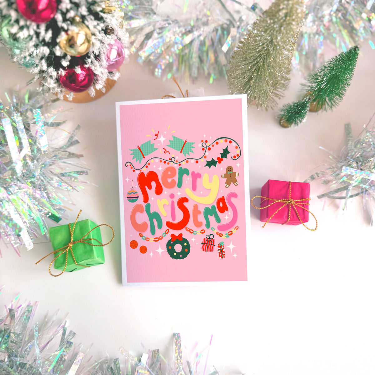 A bright pink Christmas greeting card with colourful retro-style lettering and festive illustrations, styled on a light surface with silver tinsel, miniature wrapped gifts, and holiday decorations.