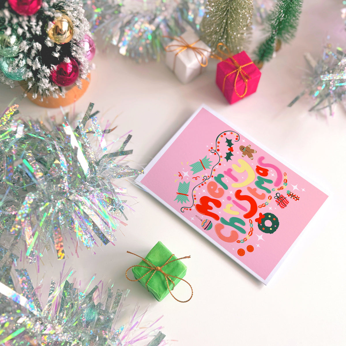 A bright pink Christmas greeting card with colourful retro-style lettering and festive illustrations, styled on a light surface with silver tinsel, miniature wrapped gifts, and holiday decorations.