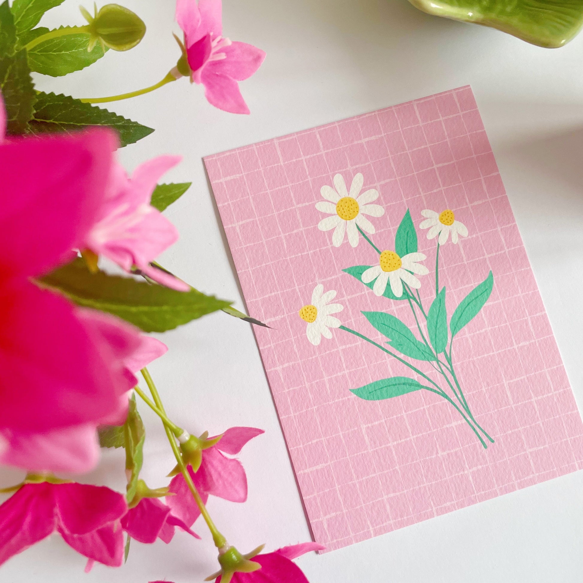 A pink textured postcard with an illustration of four daisies with bright green stems.