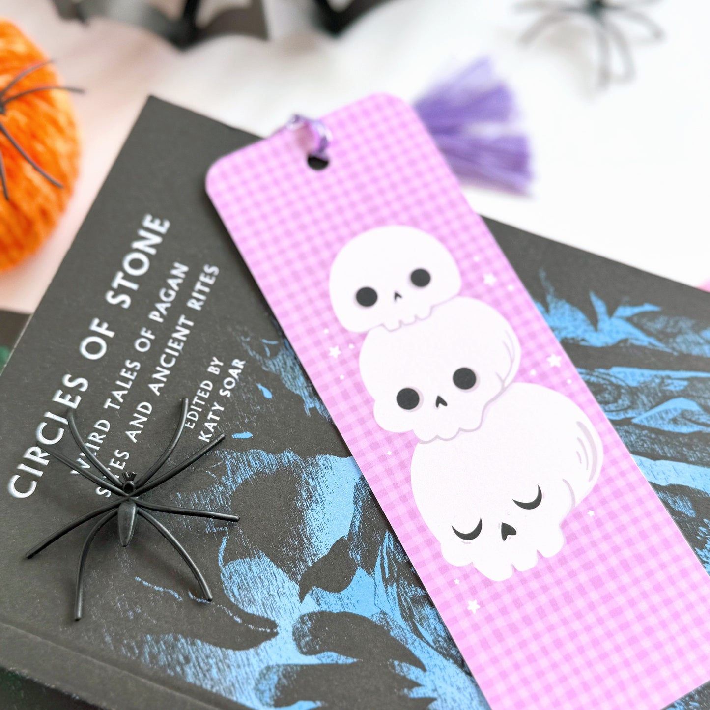 A purple gingham patterned bookmark, with a skull illustration on the front. On the back are pumpkins and the brand logo. The bookmark has a purple tassel attached. 