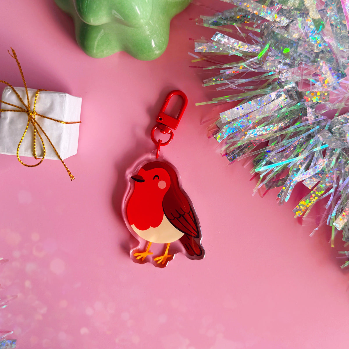 A colourful acrylic robin keychain with a red clasp. The keychain is displayed on a pink background with decorative props including a wrapped gift box, a green ceramic object, and holographic tinsel.