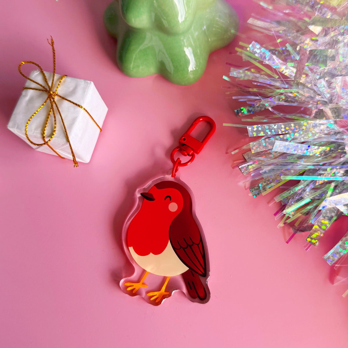 A colourful acrylic robin keychain with a red clasp. The keychain is displayed on a pink background with decorative props including a wrapped gift box, a green ceramic object, and holographic tinsel.