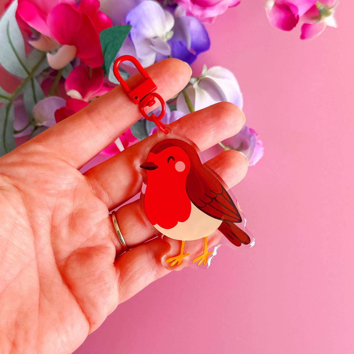 A hand holding a small bird-shaped keychain charm with a red clasp, set against a pink background with blurred flowers.