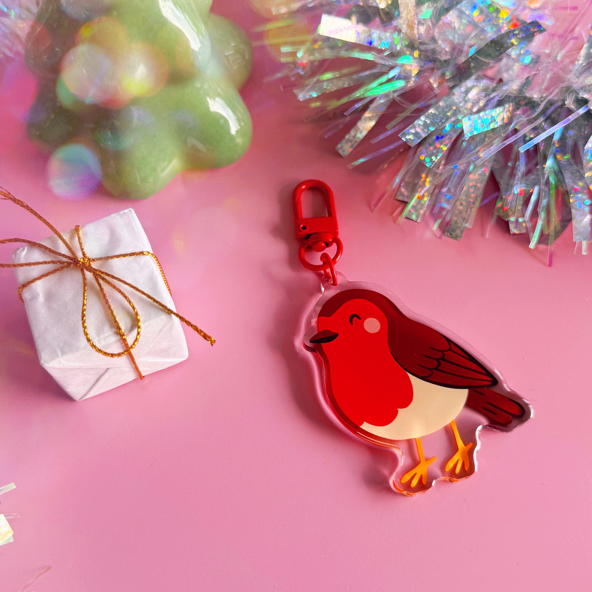 A colourful acrylic bird keychain featuring a red robin design with a red clasp, styled on a pink background with tinsel and a small wrapped gift.
